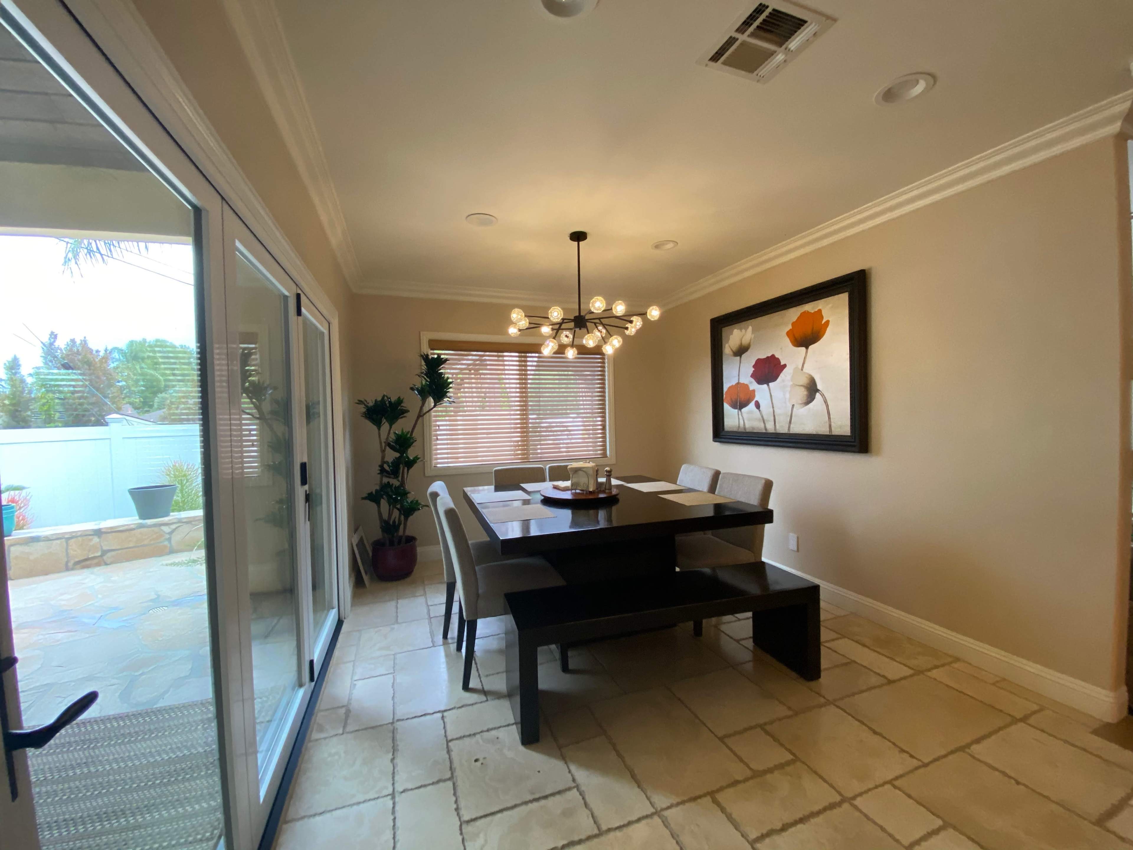 A dining area features a rectangular table surrounded by six chairs, a chandelier overhead, and a large framed artwork of flowers on the wall.