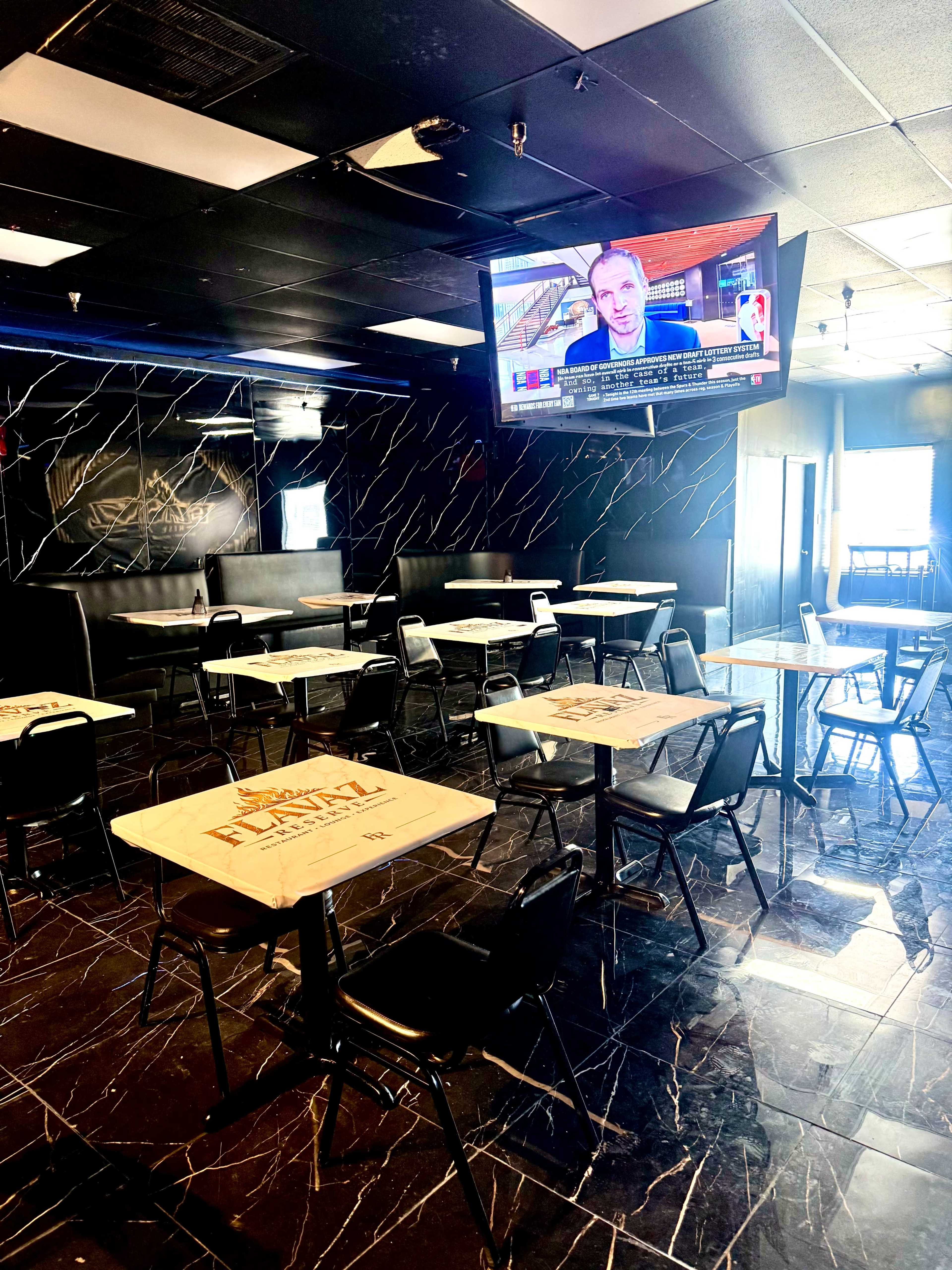 The image shows a modern, empty dining area with black chairs and tables, featuring a large television mounted on the wall displaying a news broadcast.