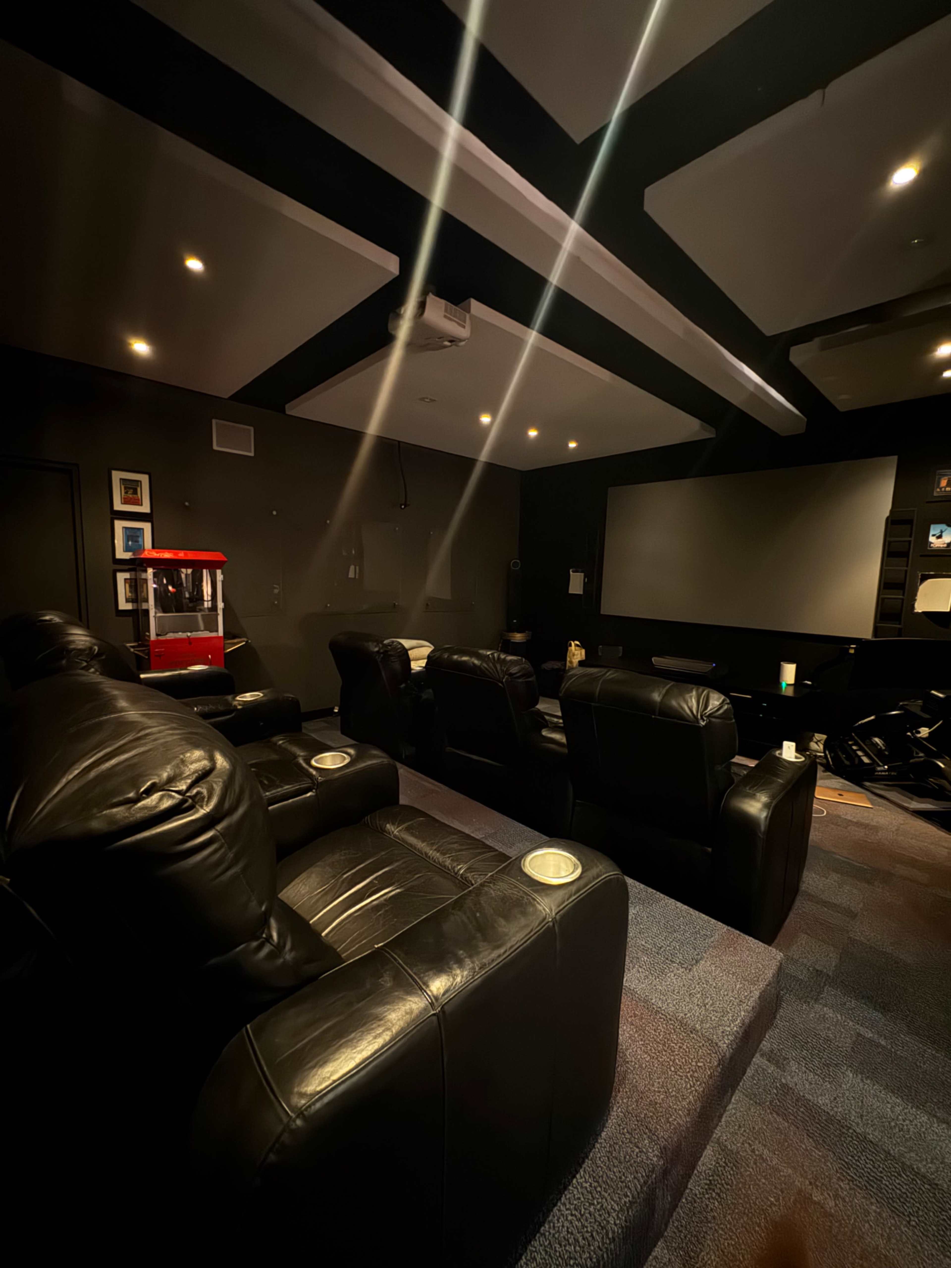The image shows a dimly lit home theater with black leather seating, a large screen in front, and a red popcorn machine in the corner.