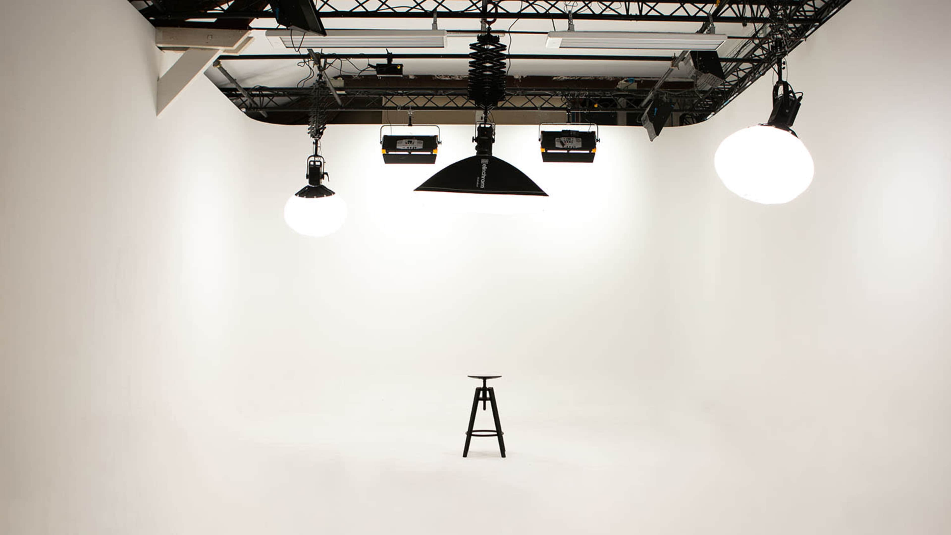 A minimalistic photography studio features a single black stool illuminated by overhead lights in a white room.