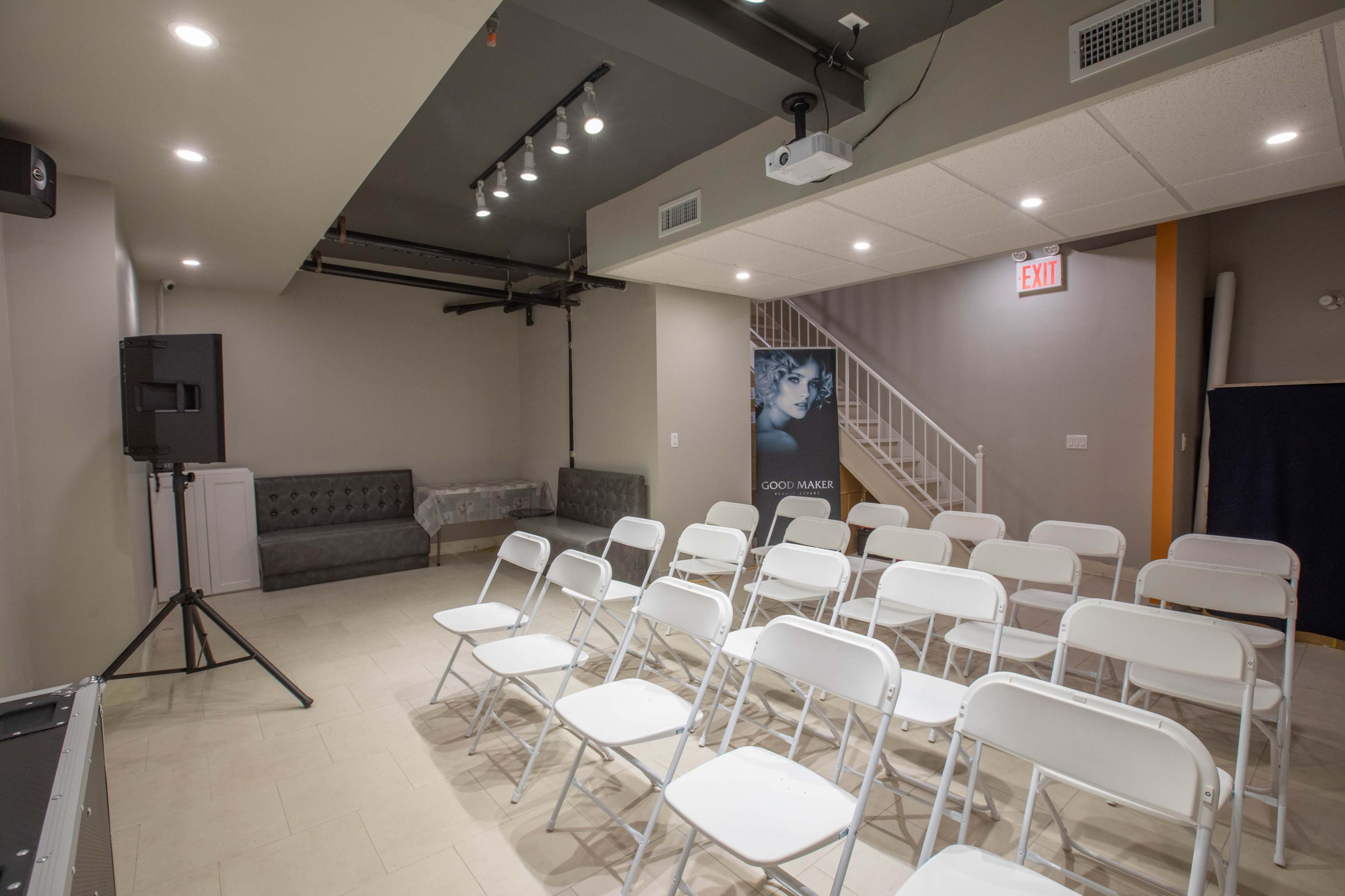 A minimalistic indoor space with white folding chairs arranged in rows facing a wall, near a gray couch and a staircase, with a promotional banner in one corner.