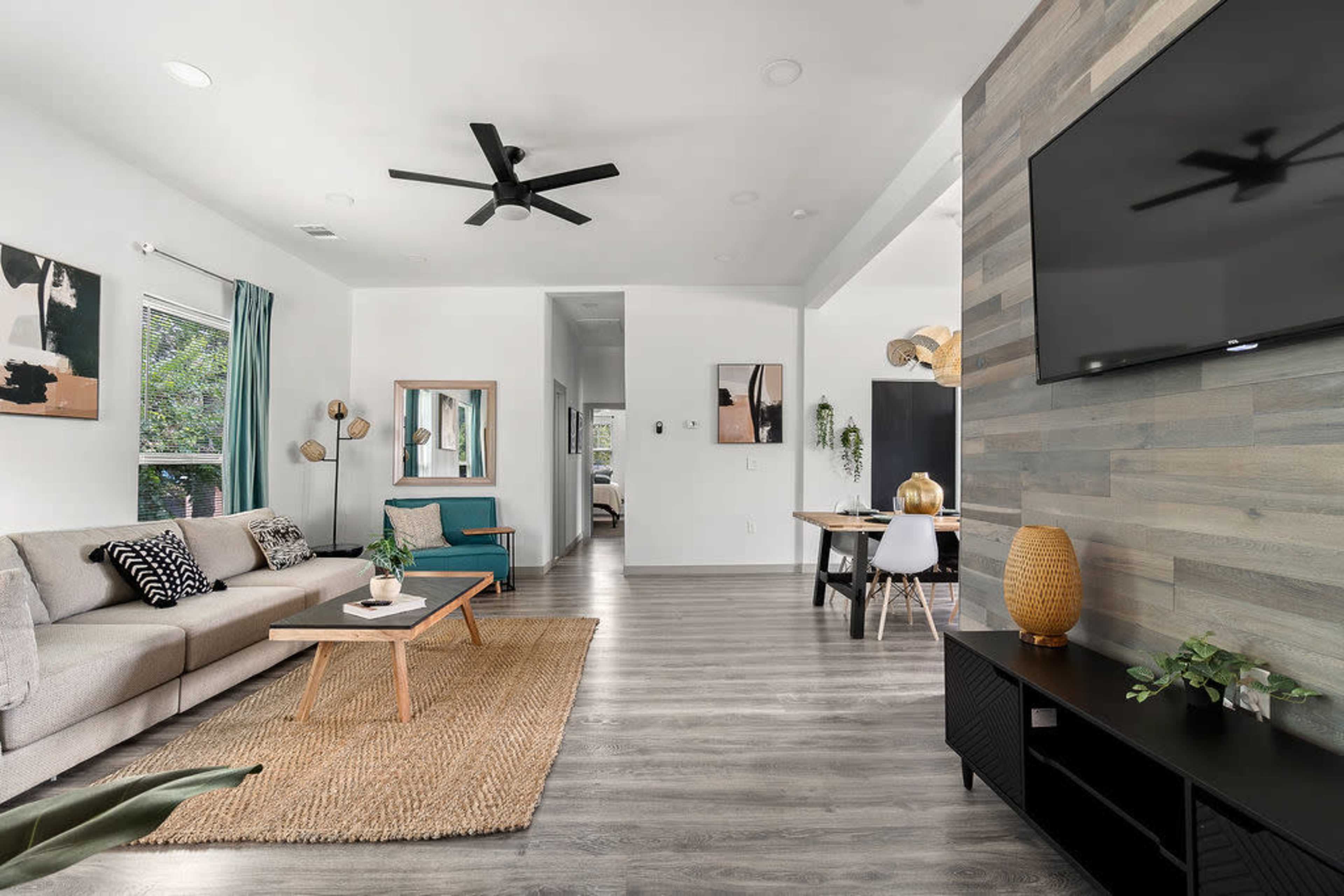 A modern living room with a gray sofa, a teal armchair, and a wooden dining area in the background, featuring a large flat-screen TV mounted on a textured wall.