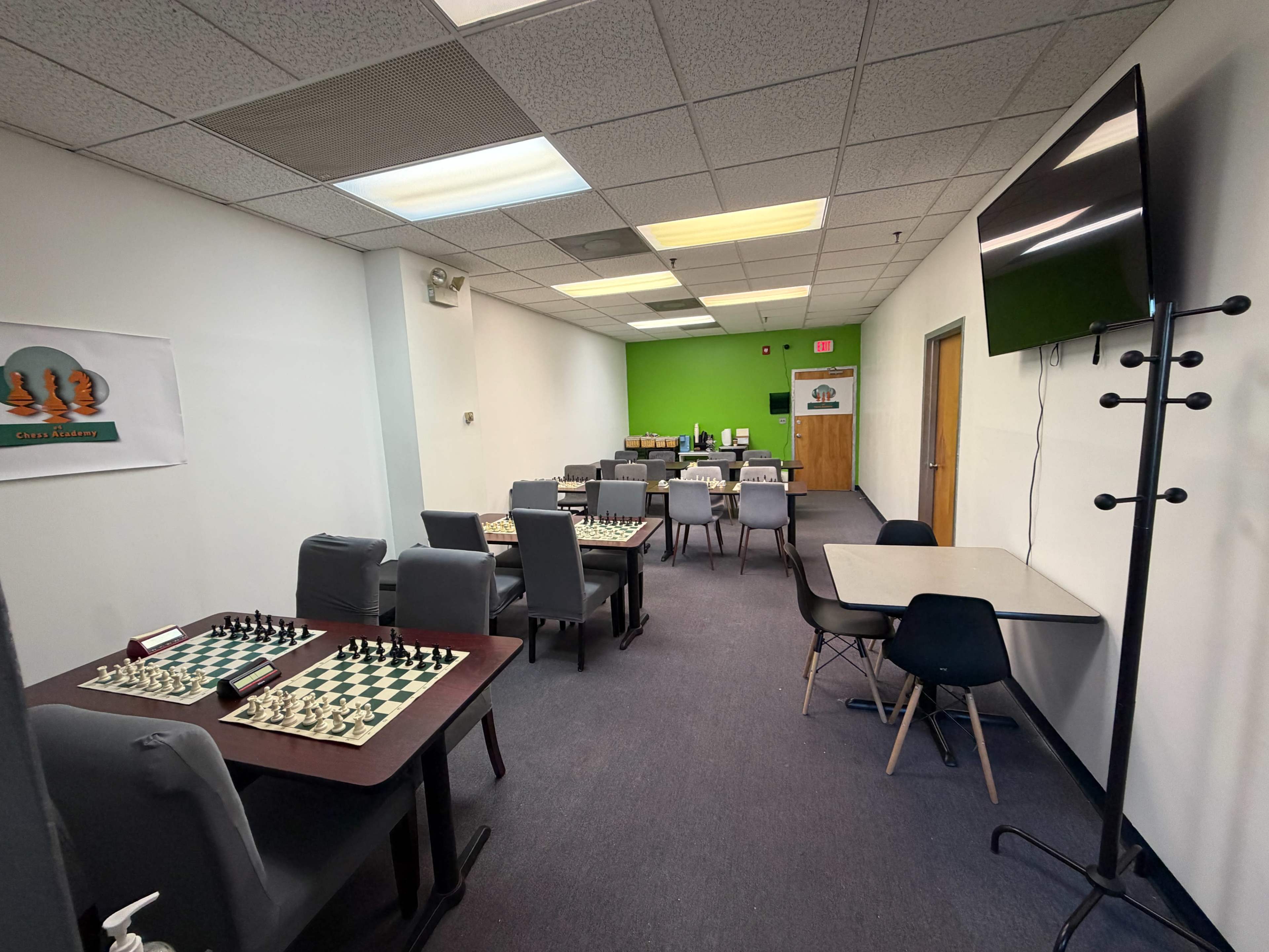 The image shows a spacious room set up for playing chess, featuring multiple tables with chessboards and chairs, along with a green accent wall and a large television.