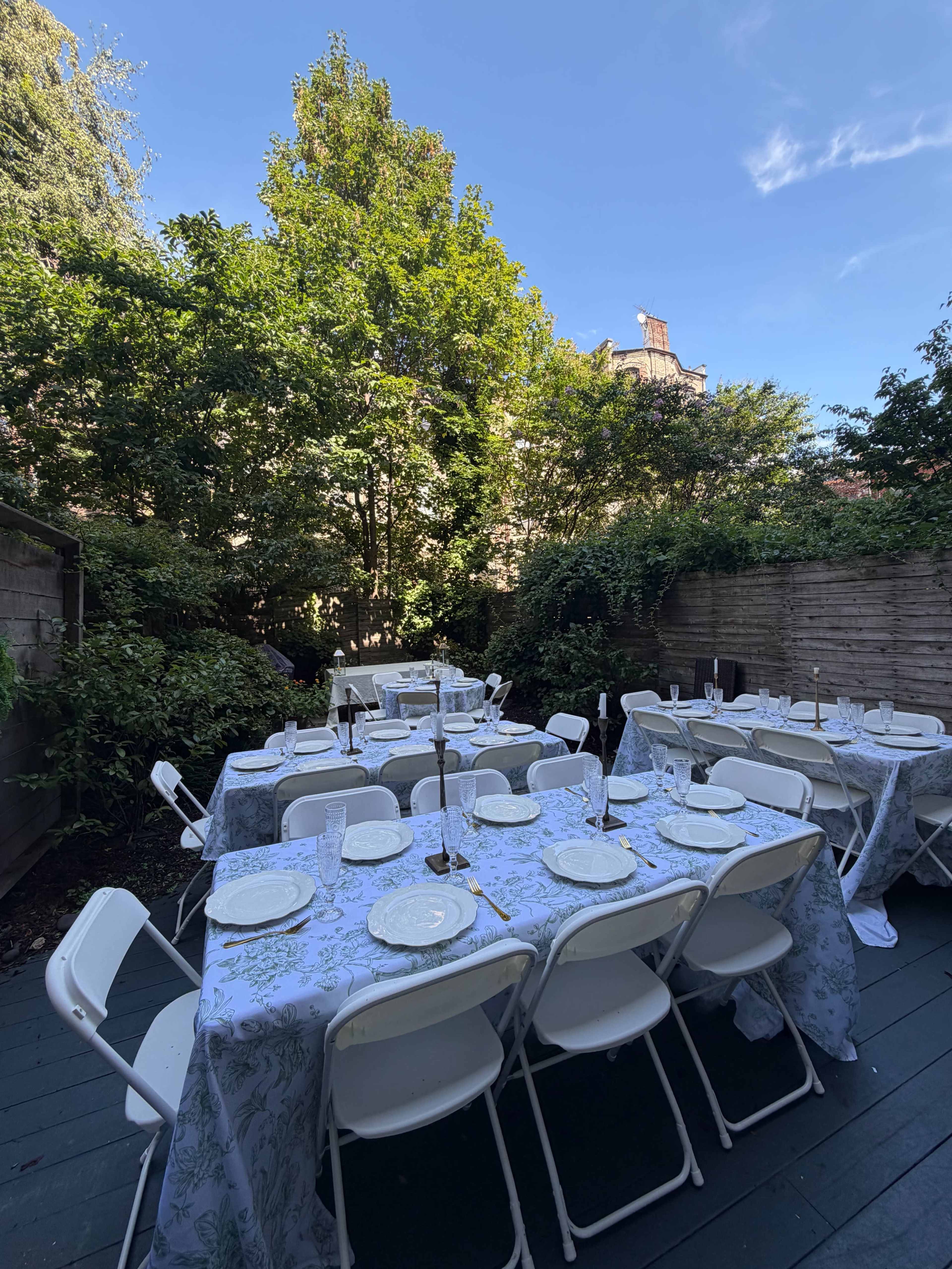 The image shows an outdoor seating area with several tables covered in white tablecloths arranged under trees in a garden setting.