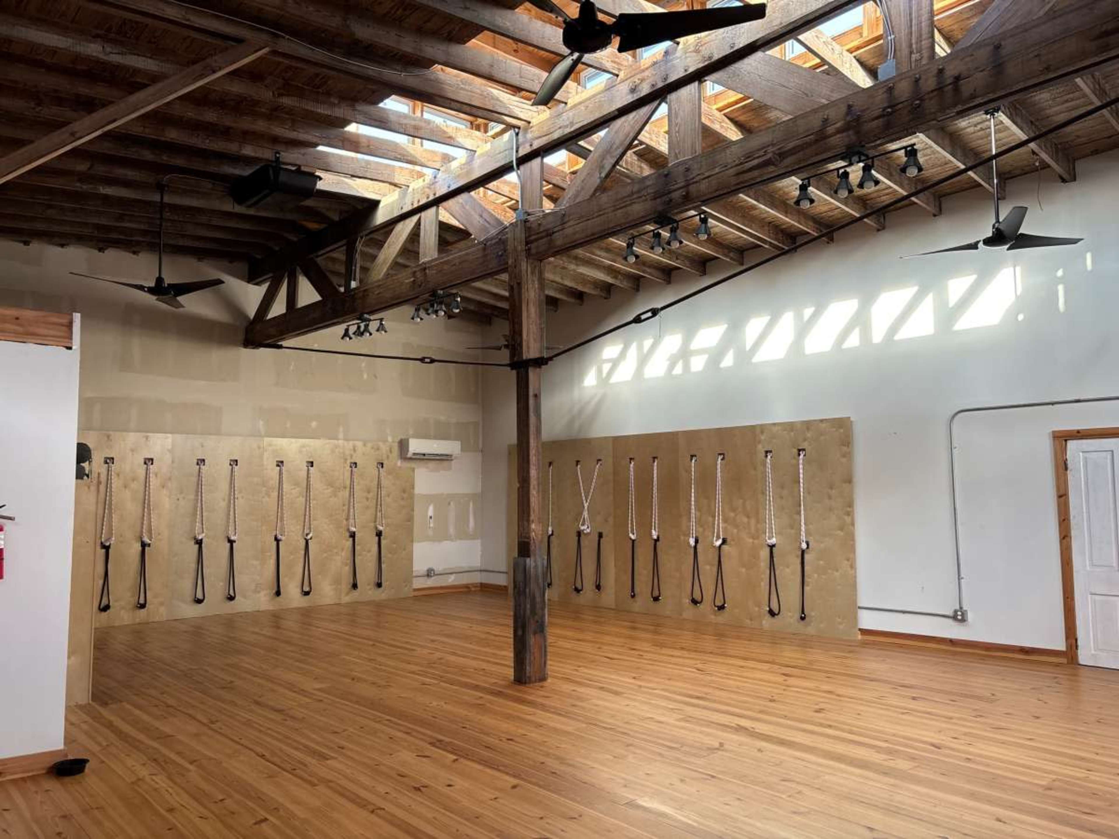 The image shows an indoor space with wooden beams and floors, featuring two walls with vertical straps attached and several ceiling fans.
