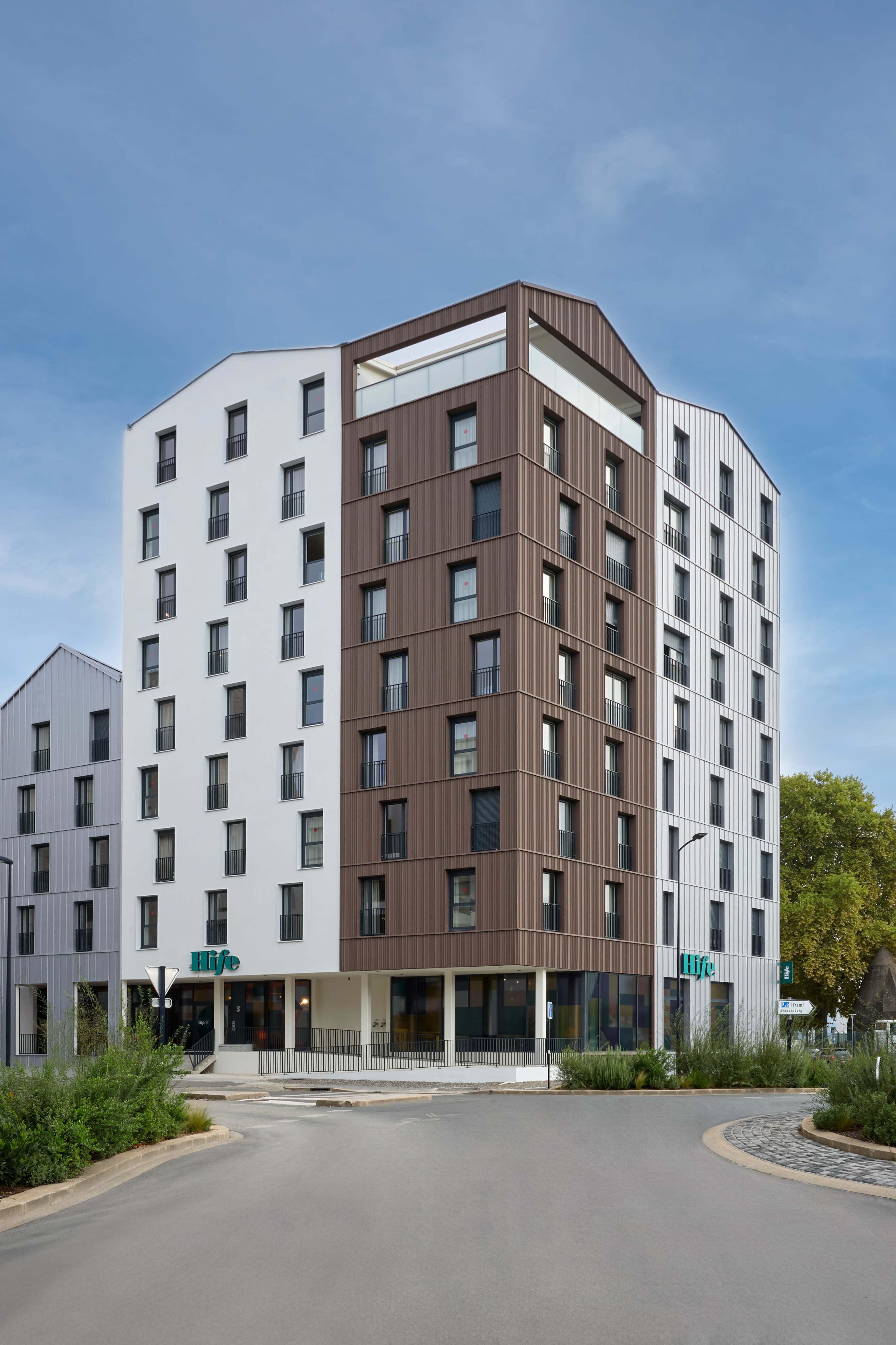 The image shows a modern, multi-story building with a geometric design featuring a combination of white and brown paneling.