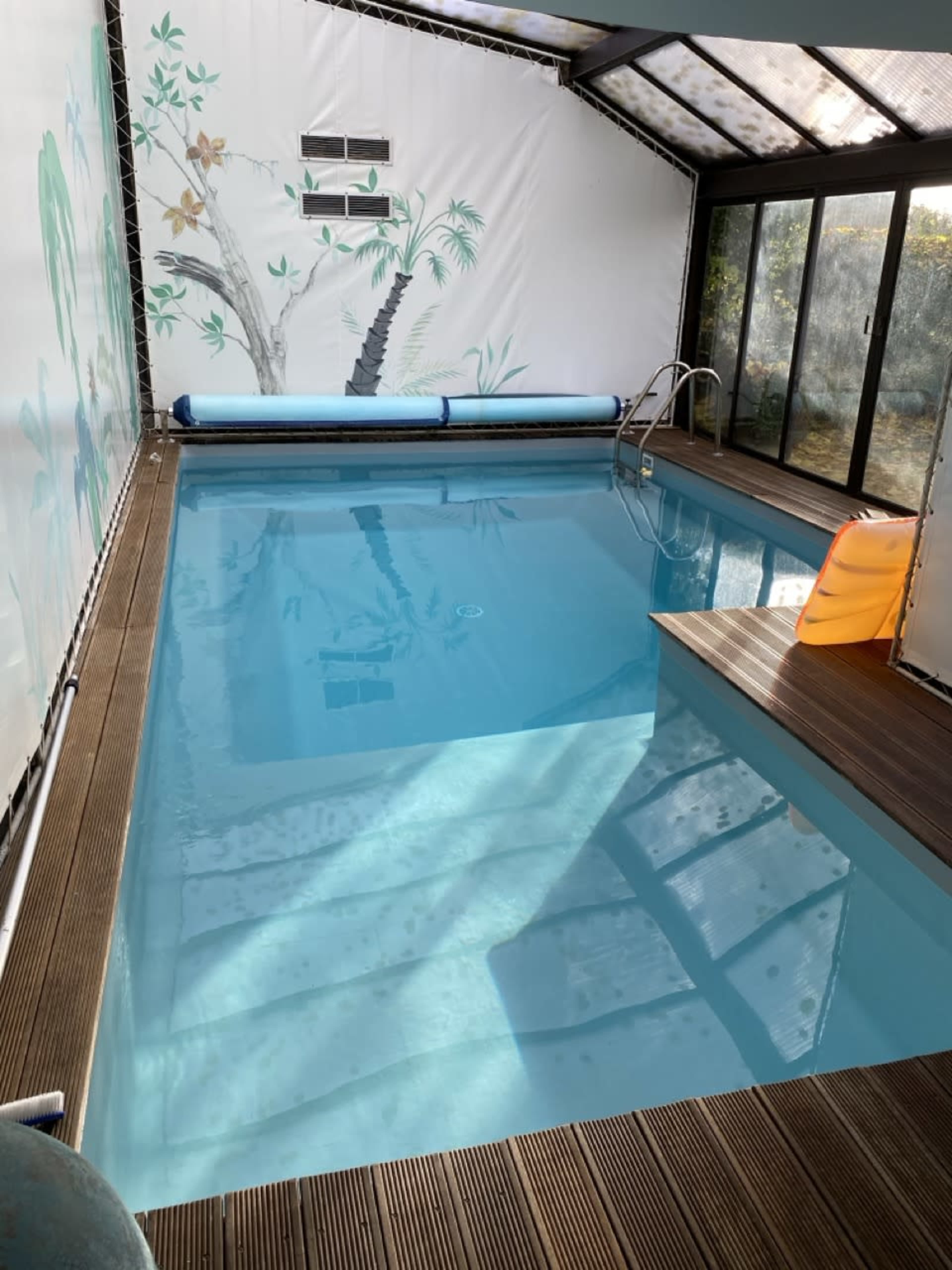 An indoor swimming pool surrounded by a wooden deck, with a mural of palm trees on one wall and a large window letting in natural light.