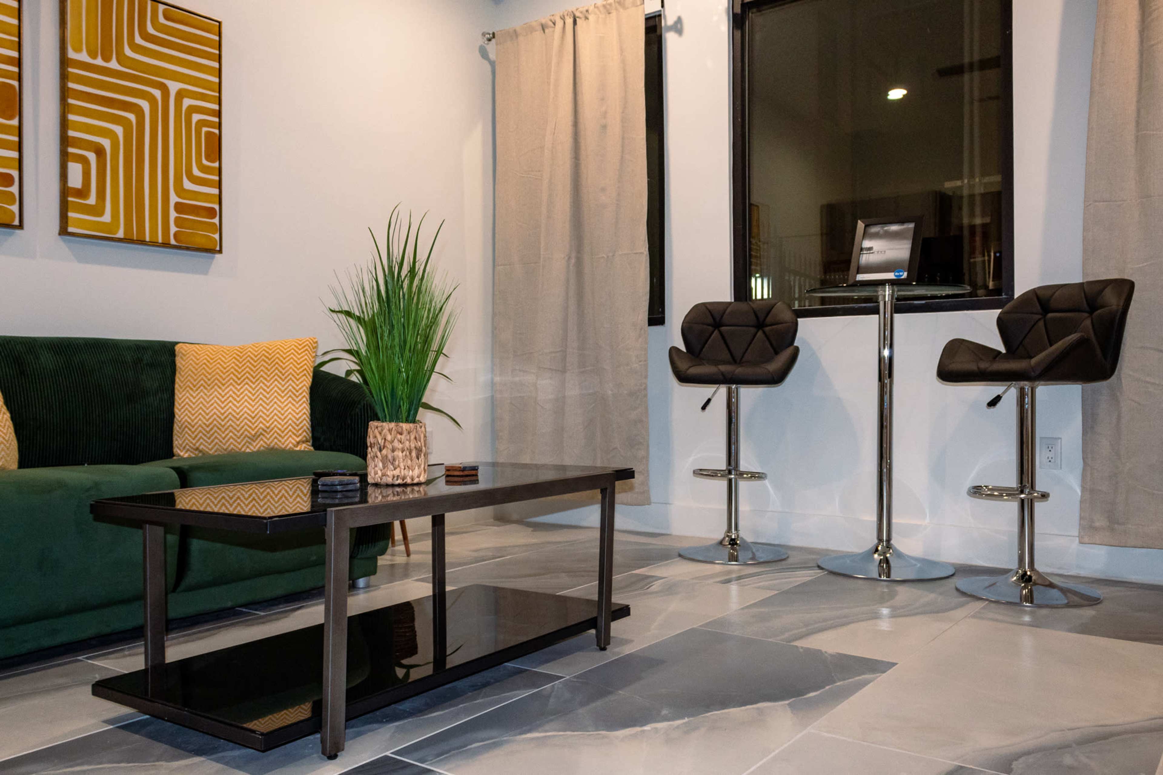 The image shows a modern living space with a green sofa, a coffee table, and two bar stools near a window.