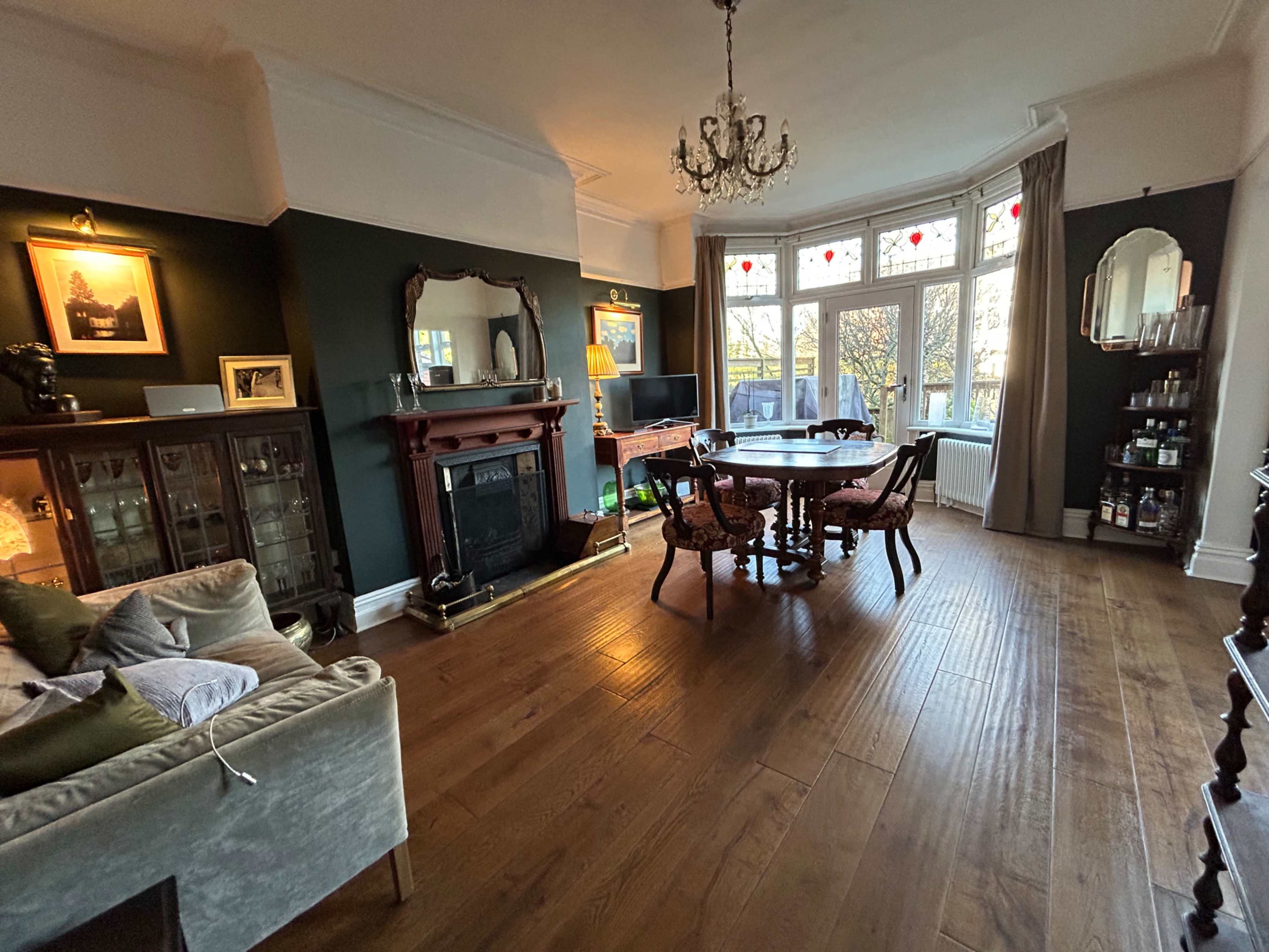 The image shows a spacious living room featuring wooden flooring, a large dining table with chairs, a fireplace adorned with a mirror, and a bay window with light streaming in.