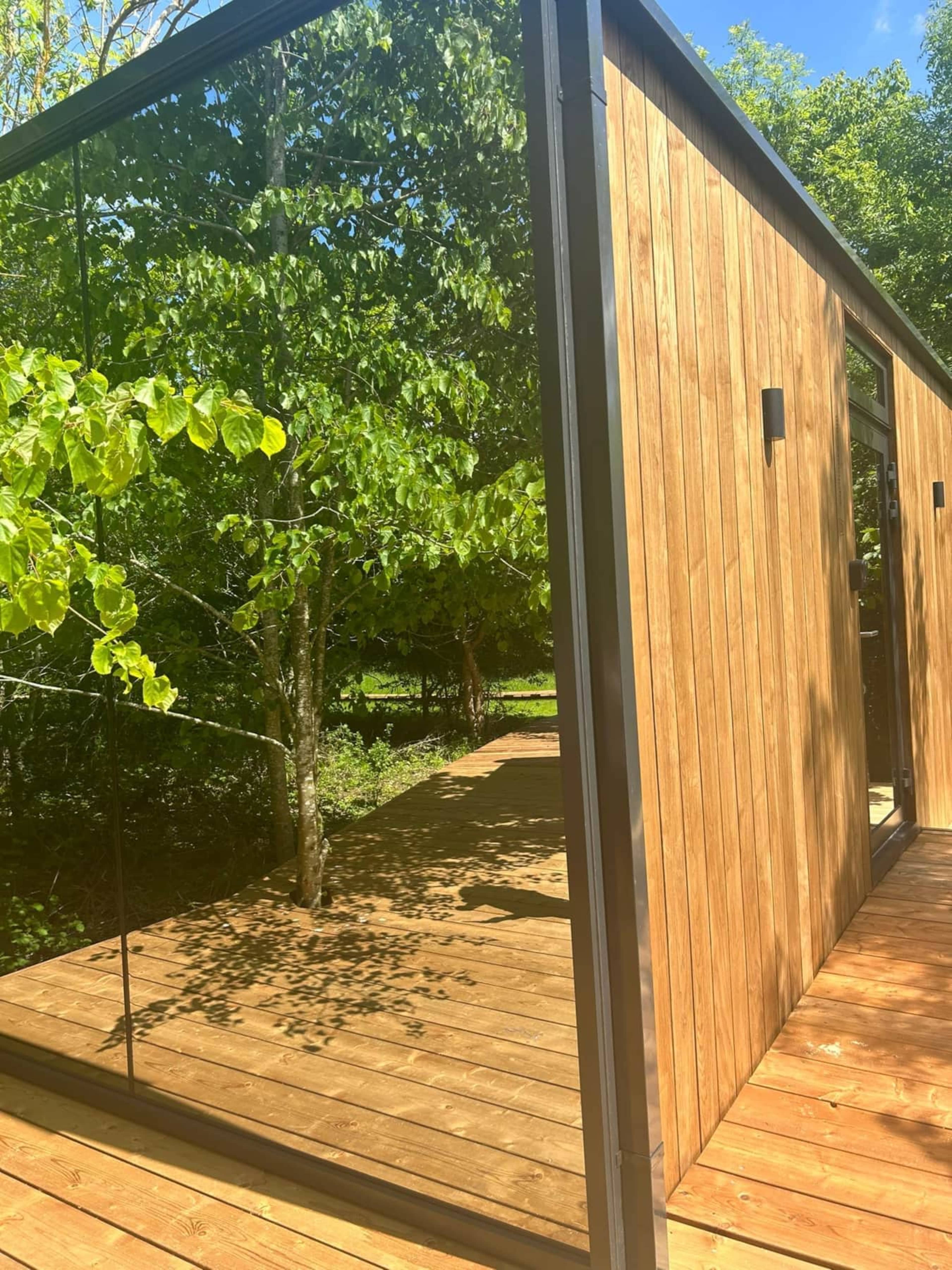 A wooden structure with a glass wall reflecting greenery, surrounded by trees and a wooden deck.