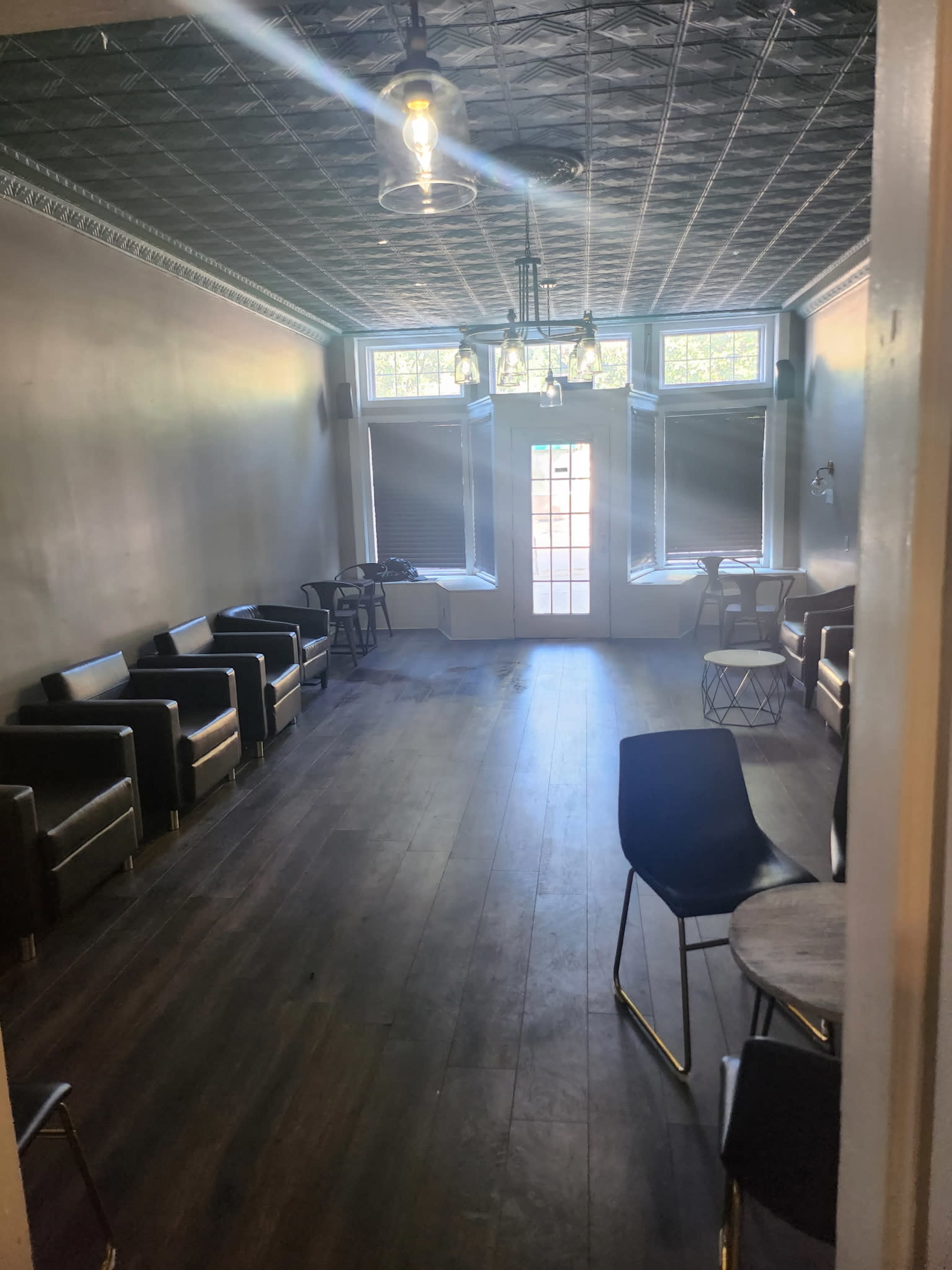 The image shows a spacious, well-lit room furnished with several chairs and tables, featuring large windows and a patterned ceiling.