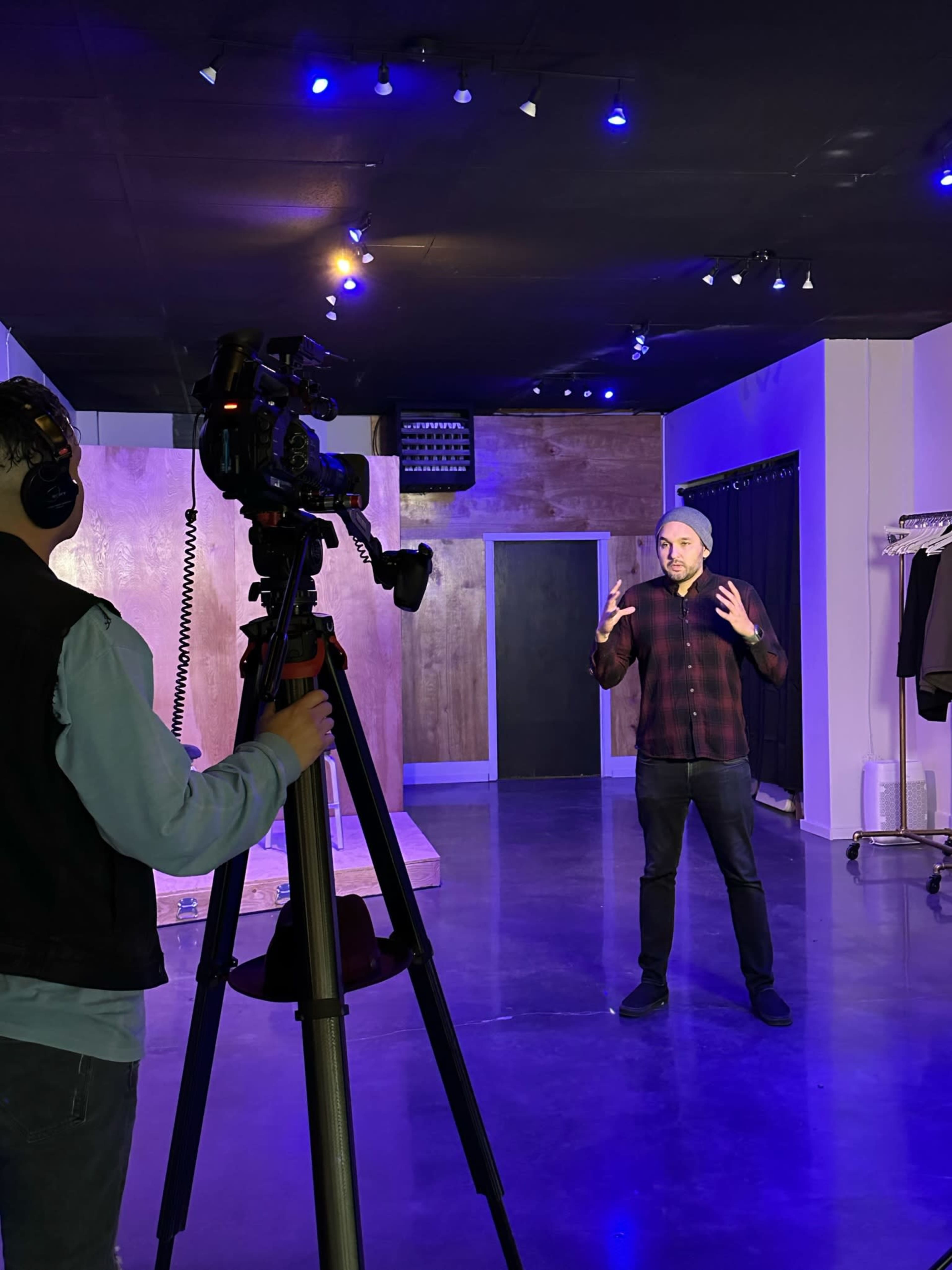 A person is filming another individual speaking in a studio with purple lighting and a minimalist backdrop.