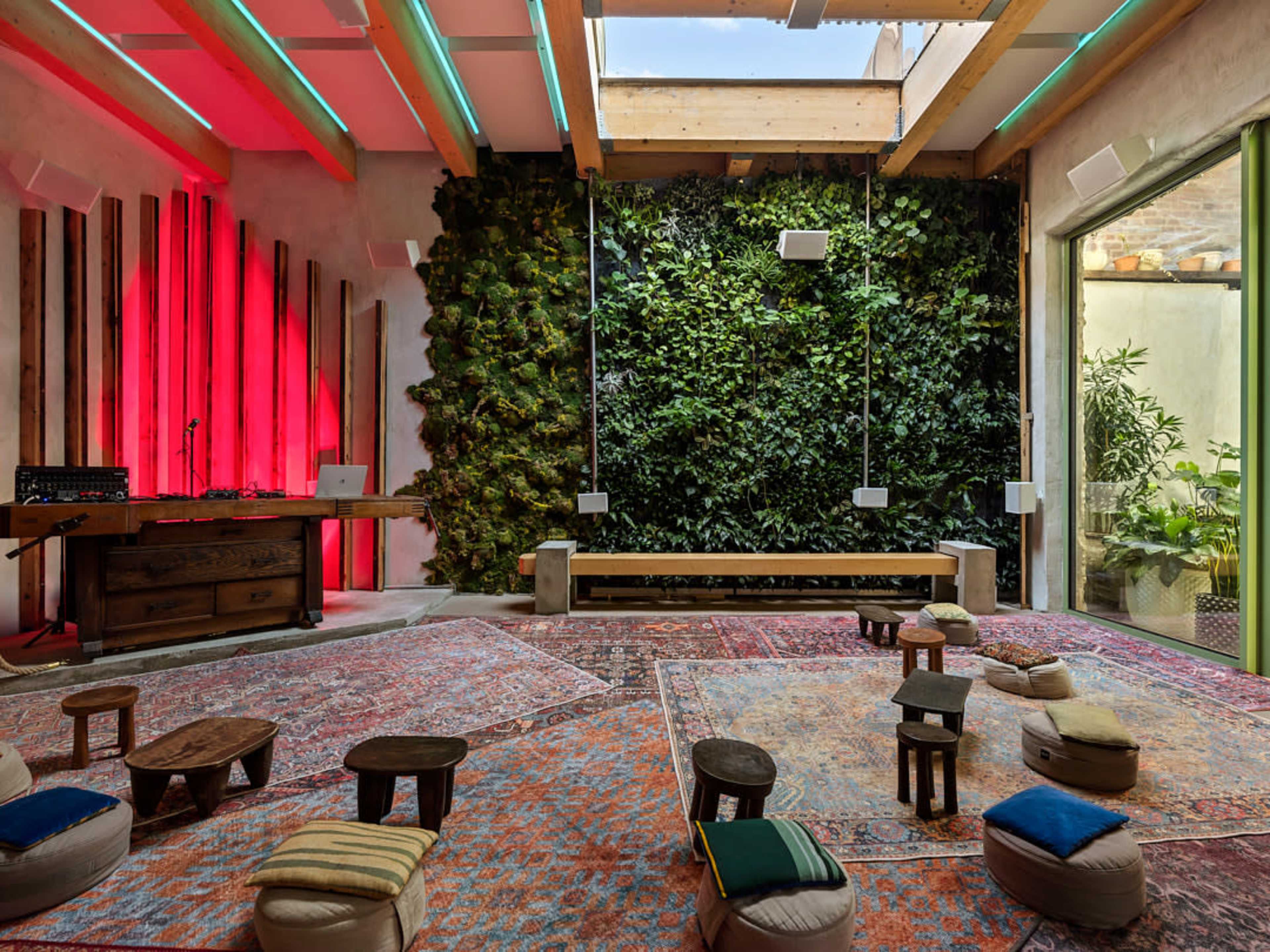 The image shows a modern, indoor space with a green living wall, seating arranged on colorful rugs, and a wooden DJ setup in the corner.