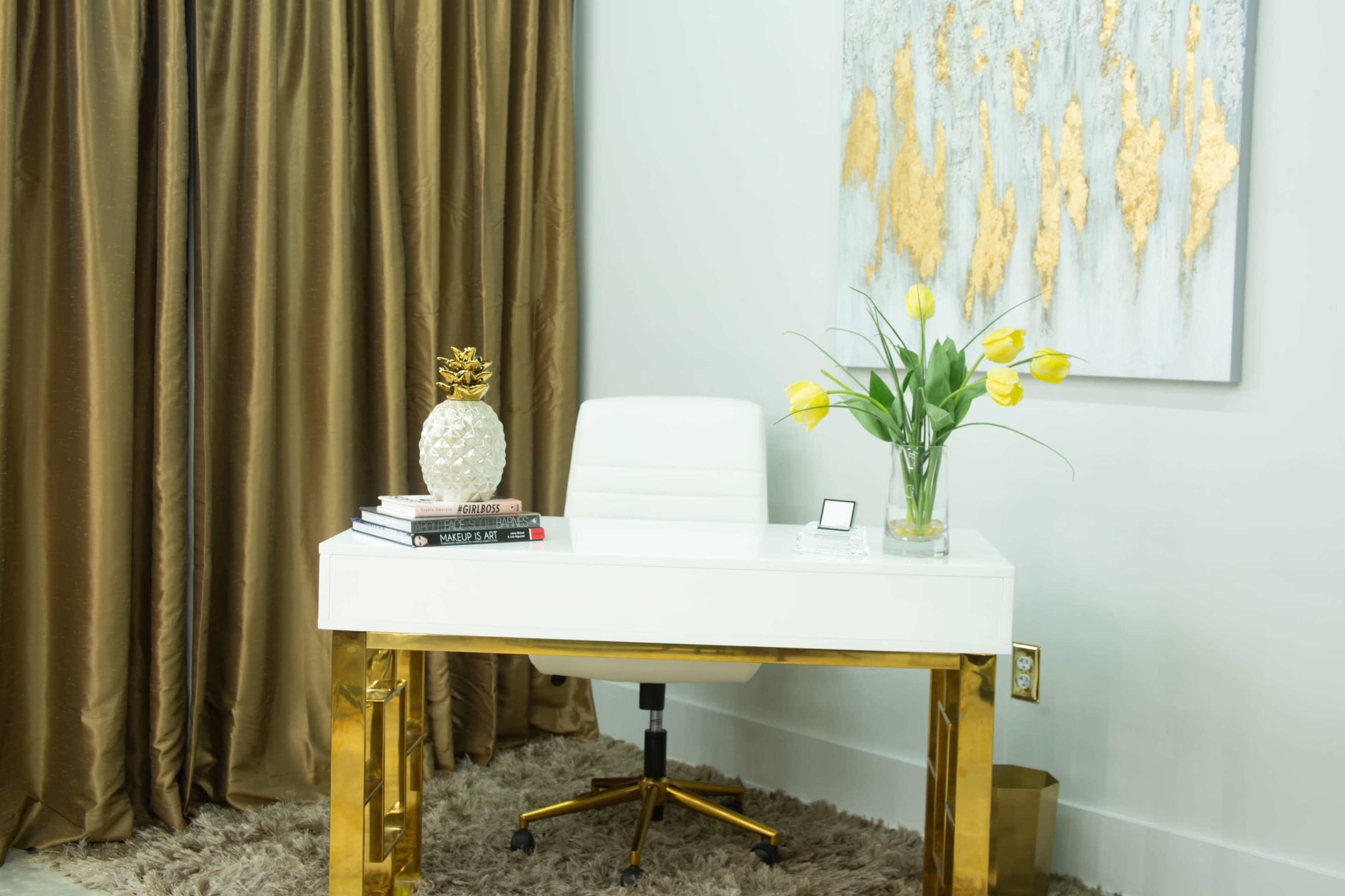 A modern desk with a white chair is positioned near a gold-accented wall, decorated with a floral centerpiece and surrounded by brown curtains.