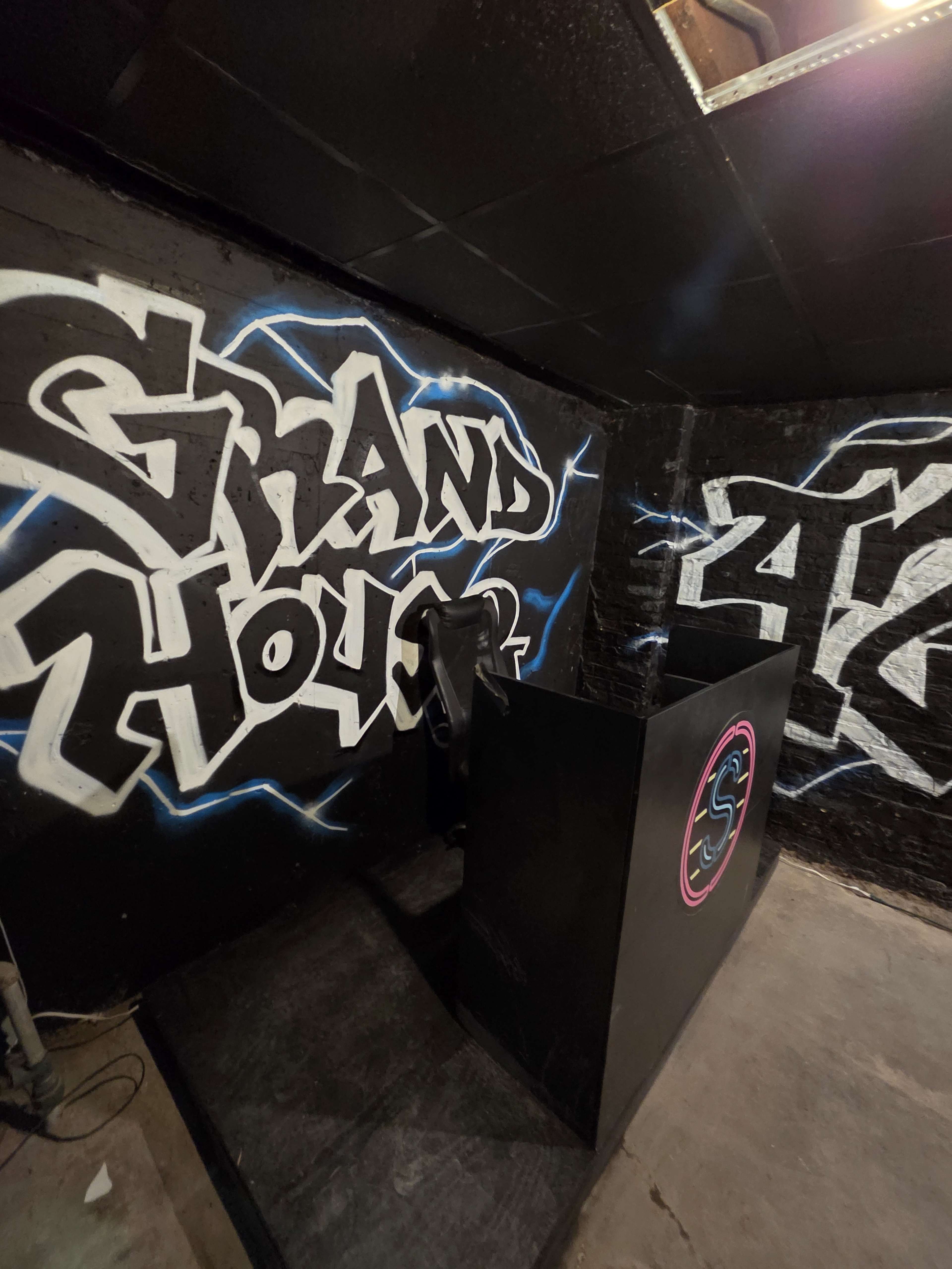 The image depicts a DJ booth situated against a wall adorned with graffiti that reads "Grand House" and abstract designs.