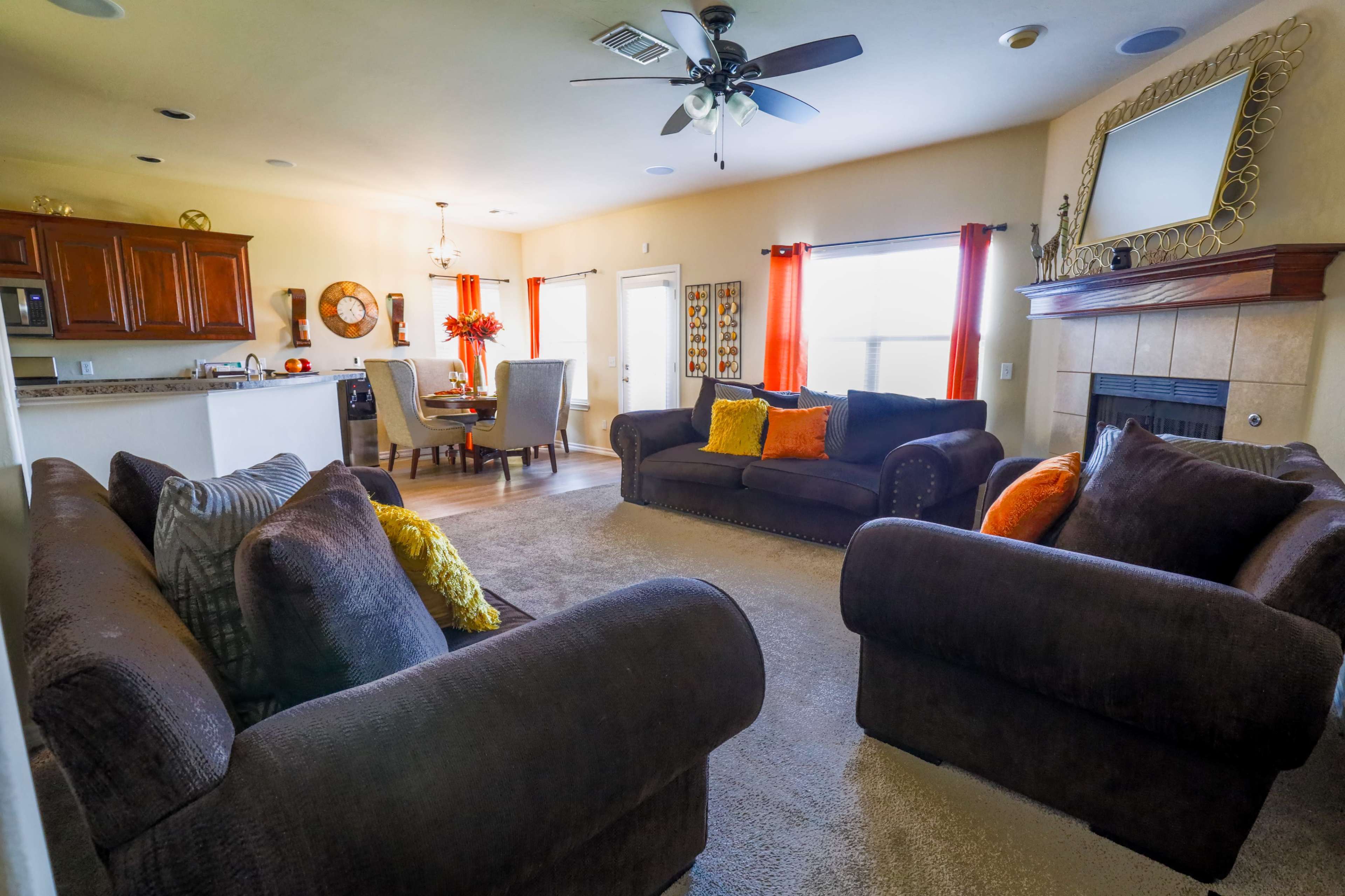 A cozy living room with two dark sofas facing each other, a round dining table with chairs, a fireplace, and large windows with orange curtains.