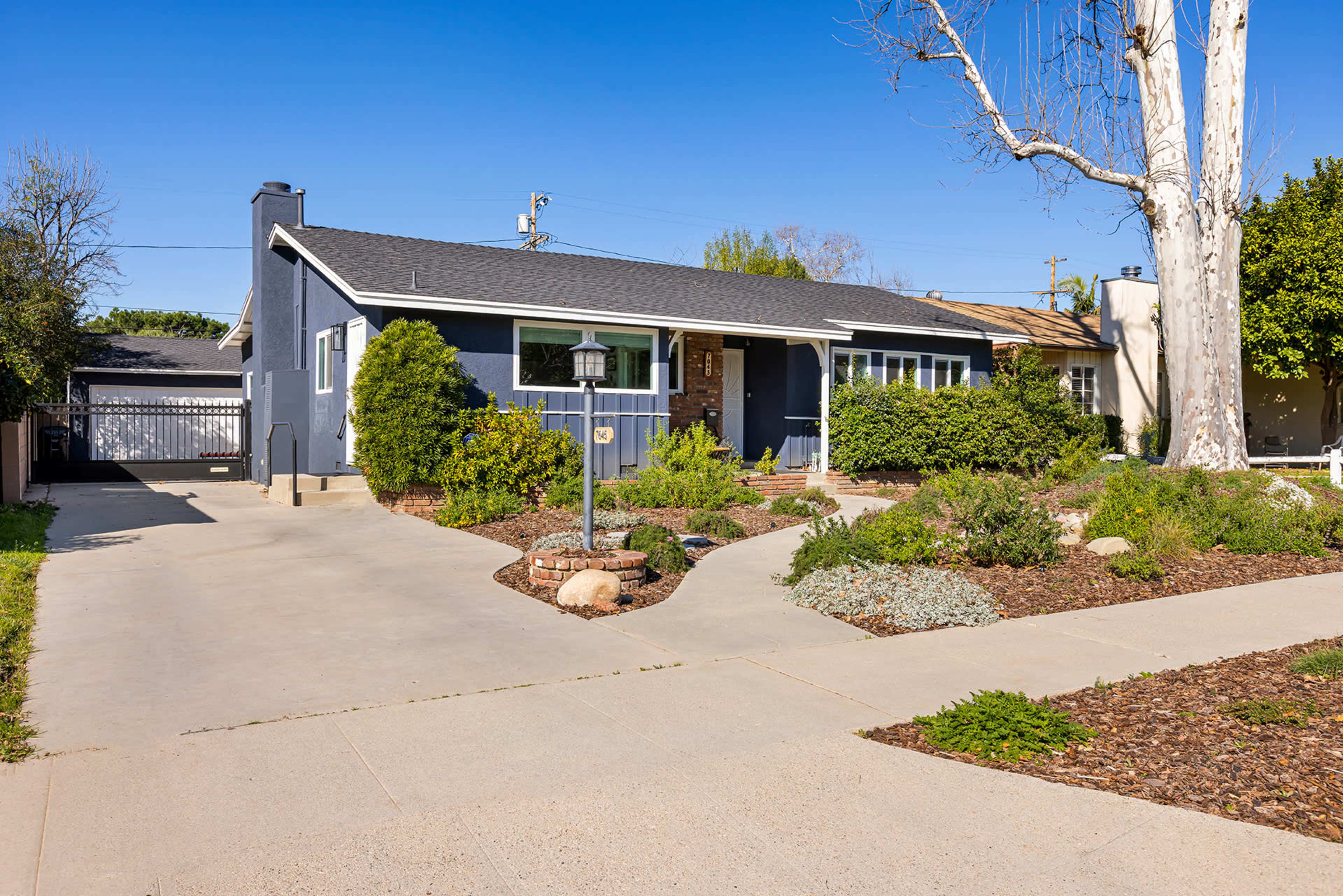 A single-story blue house with a well-maintained front yard featuring various plants and a pathway leading to the entrance.