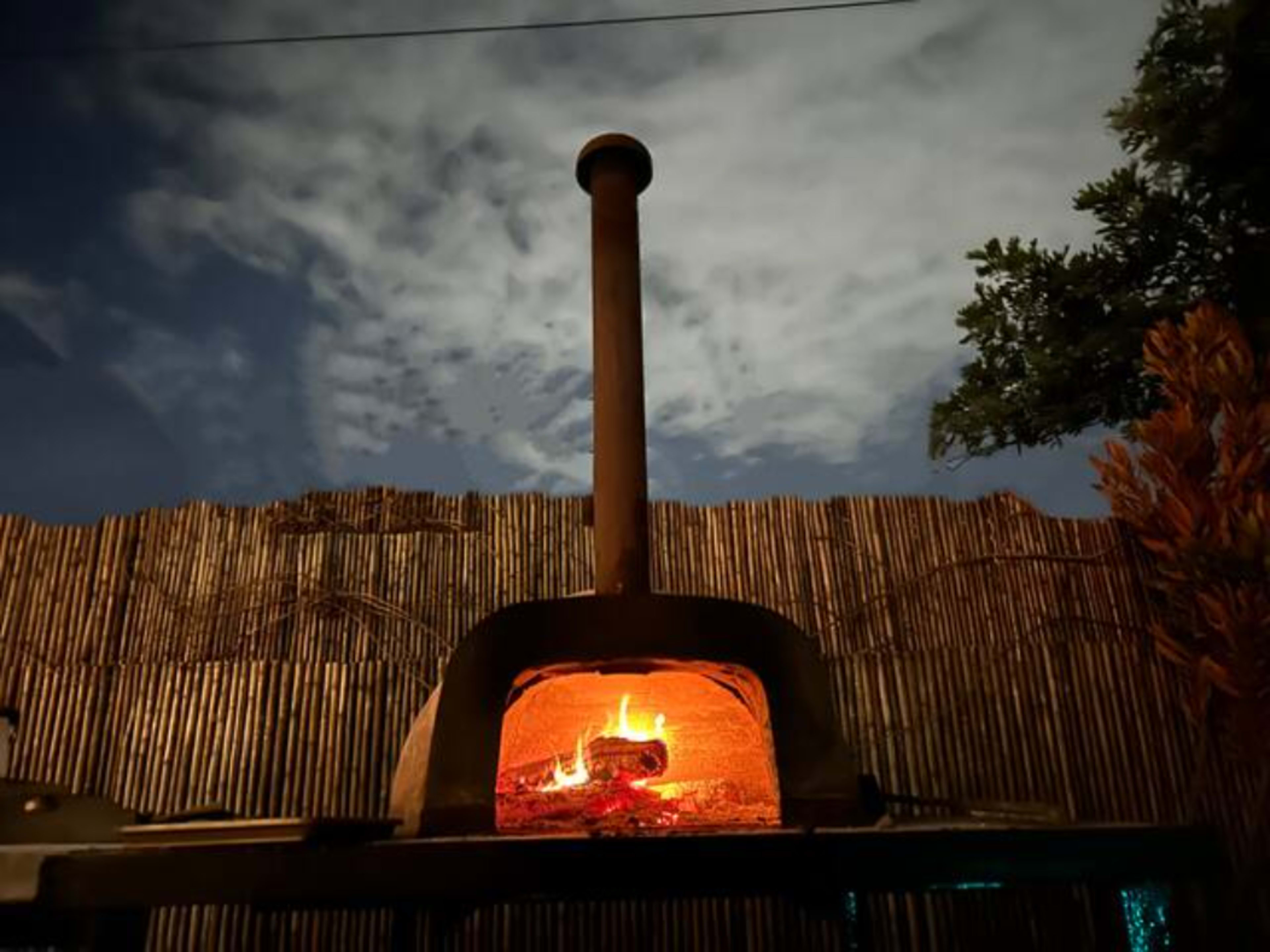 A wood-fired oven with a visible flame sits in front of a bamboo fence under a cloudy night sky.