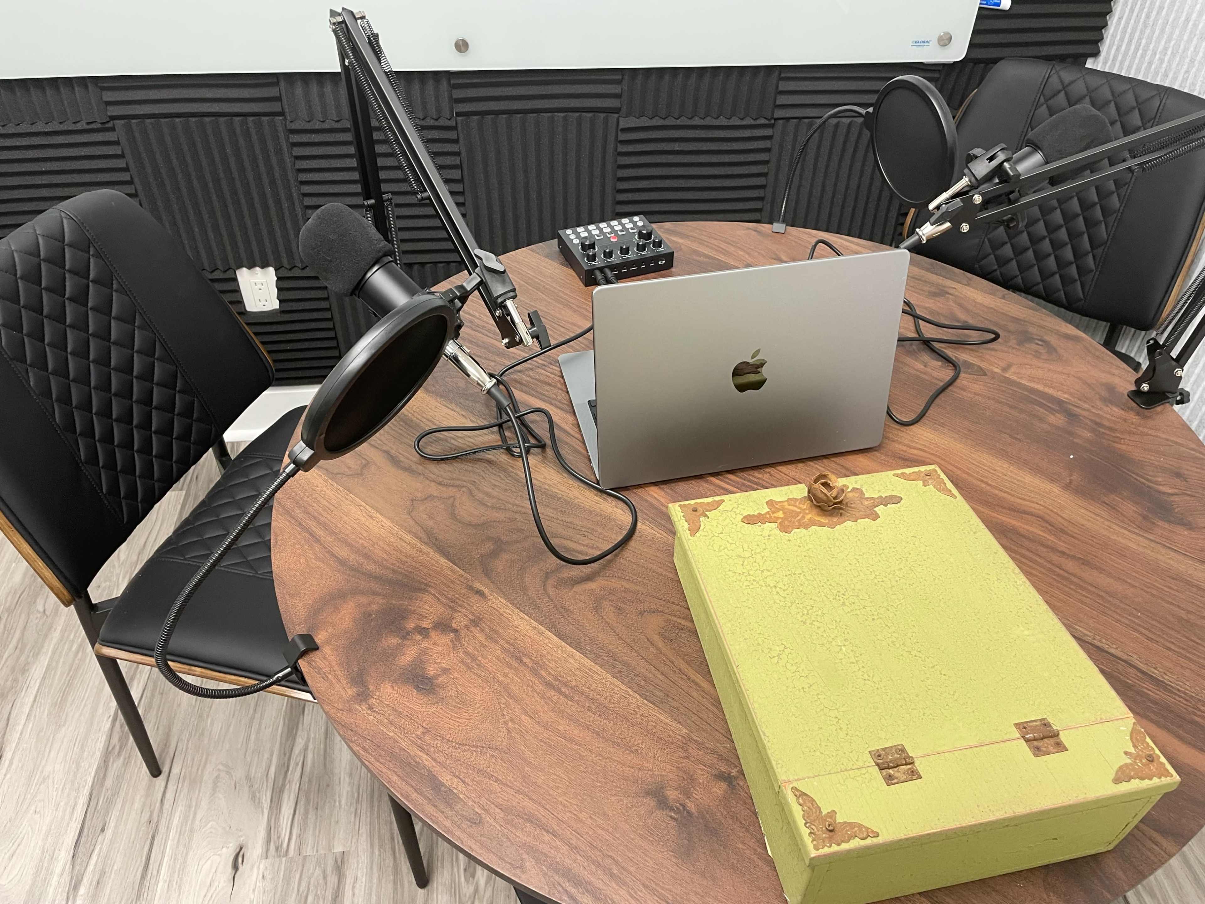 A round wooden table is set with two microphones, a laptop, and a green box in a room with soundproofing panels.