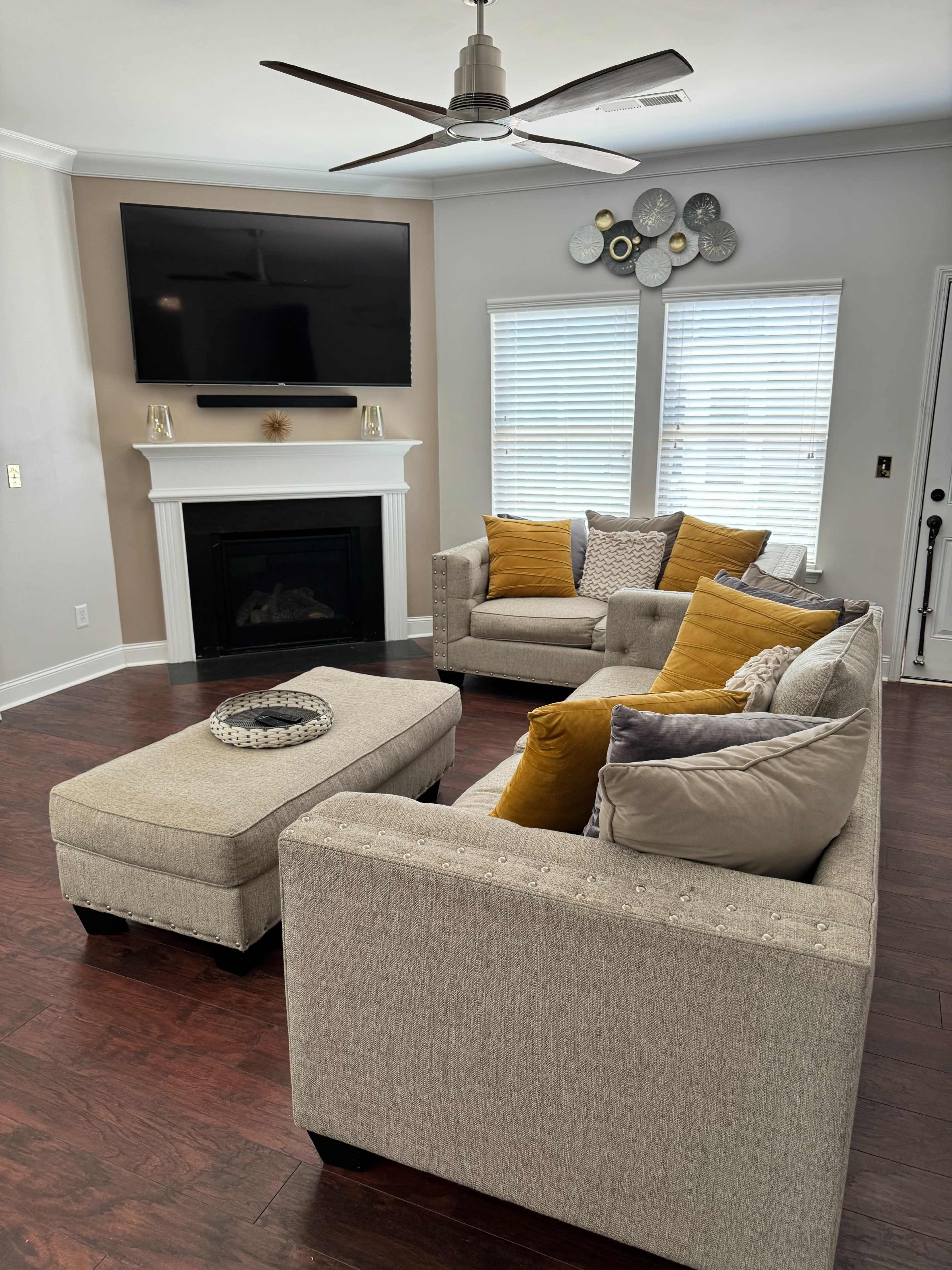 The living room features a beige sectional couch, a coffee table, an entertainment center with a flat-screen TV, and a fireplace, surrounded by large windows with blinds.