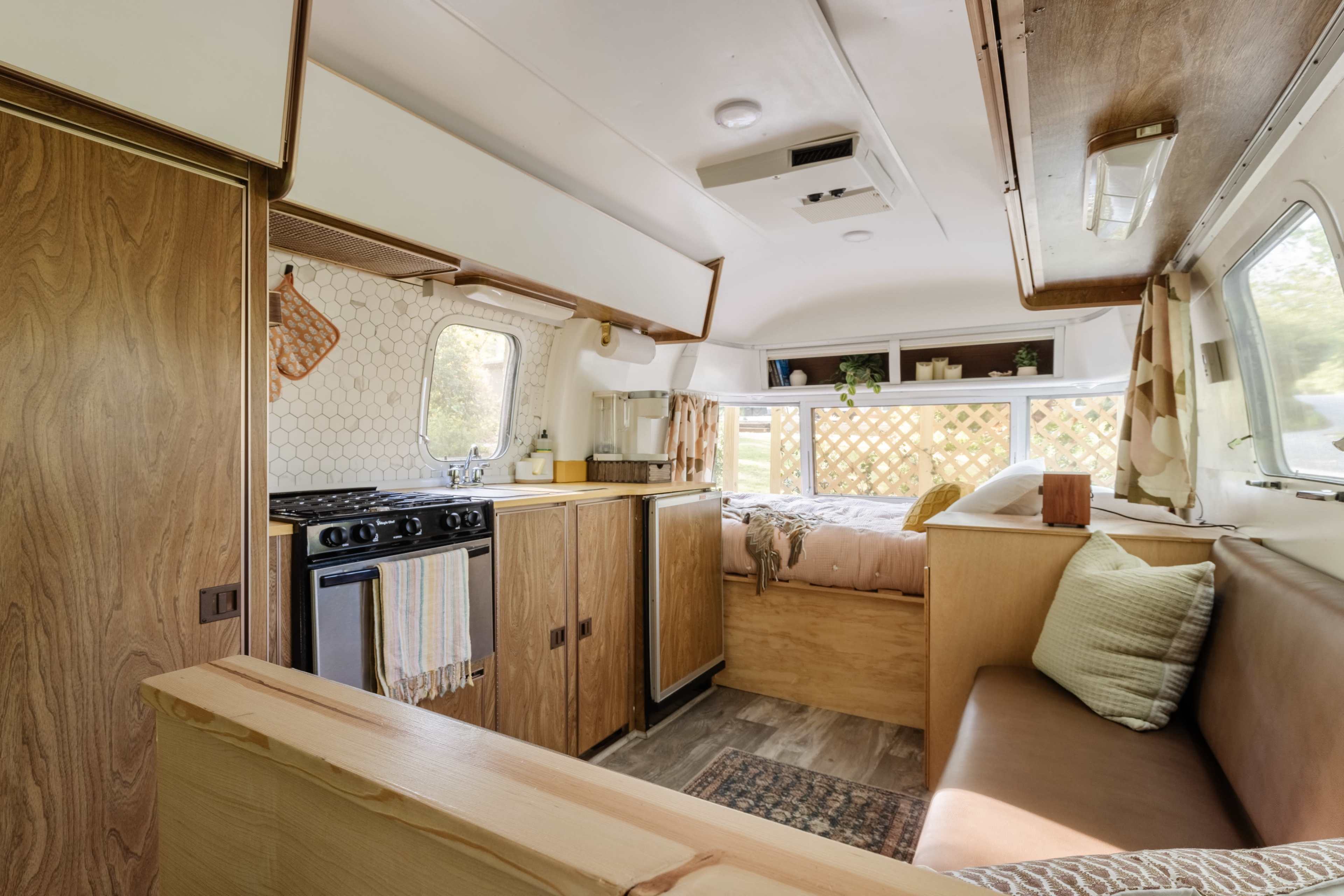 The interior of a small camper features a kitchen area with a stove and wooden cabinets, a seating nook, and a bed with a patterned throw in the rear.
