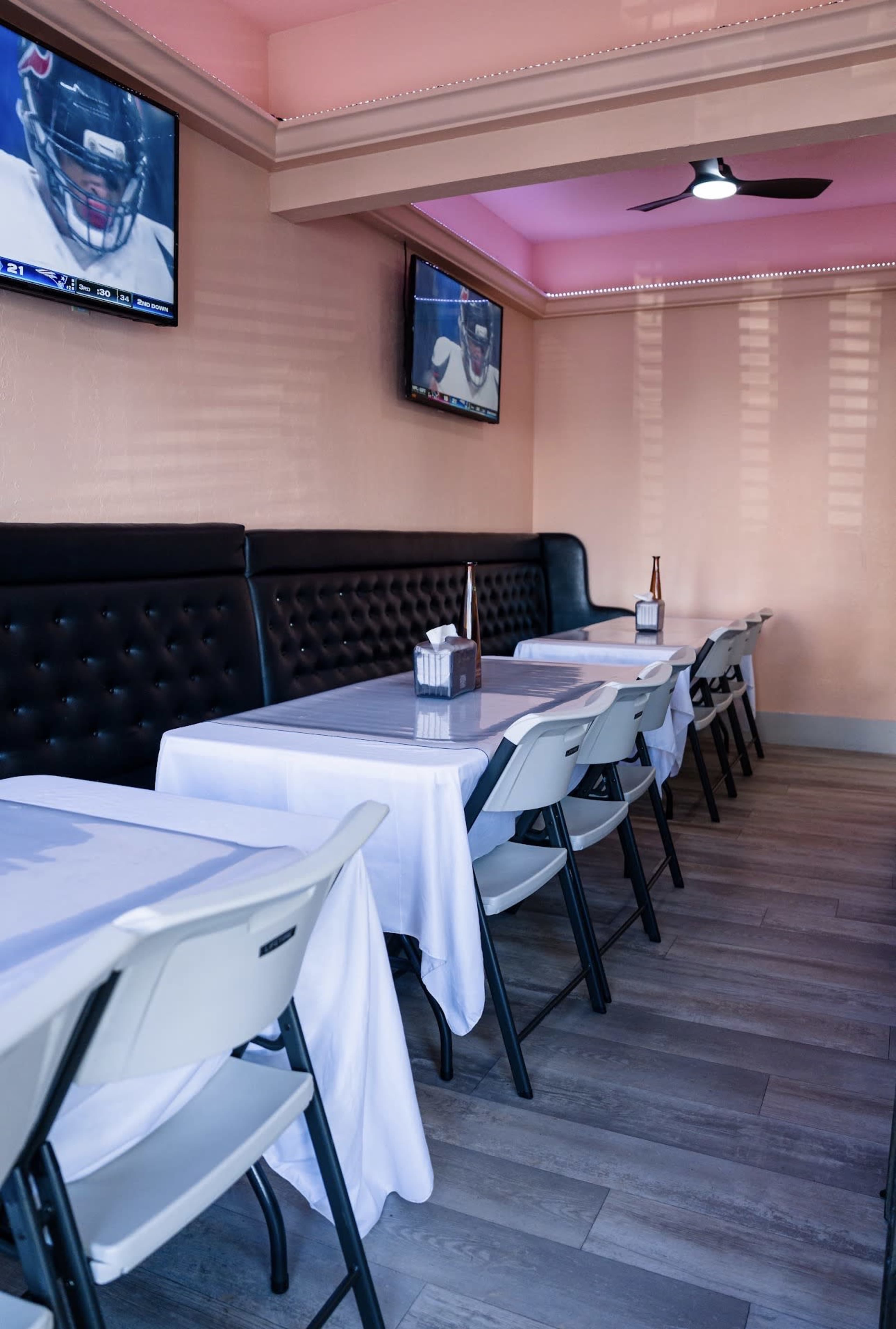 A tidy restaurant interior features several tables covered with white tablecloths, flanked by black upholstered benches, and equipped with wall-mounted televisions.