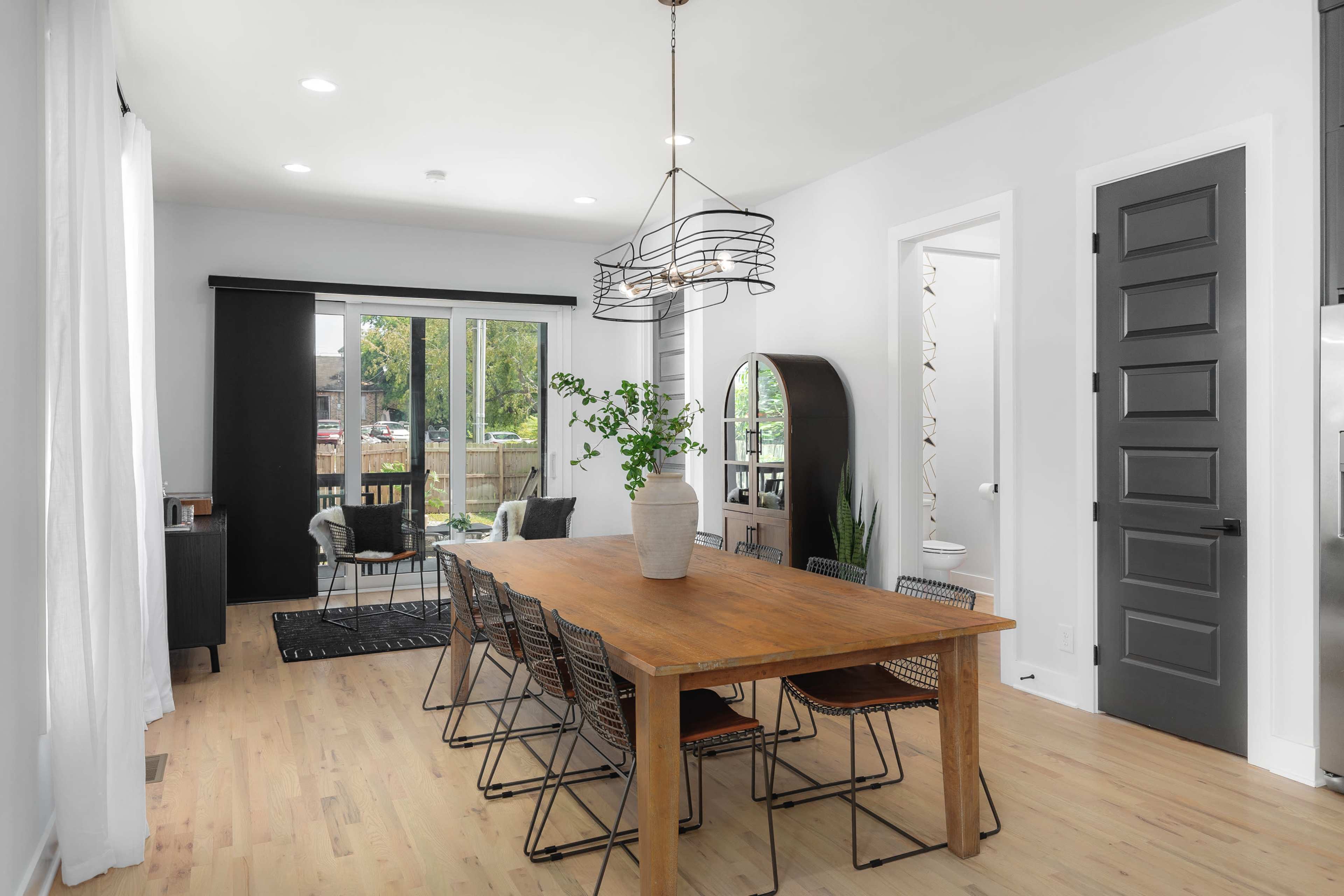 A spacious dining area features a large wooden table surrounded by metal chairs, with a modern light fixture above and sliding glass doors leading to an outdoor space.