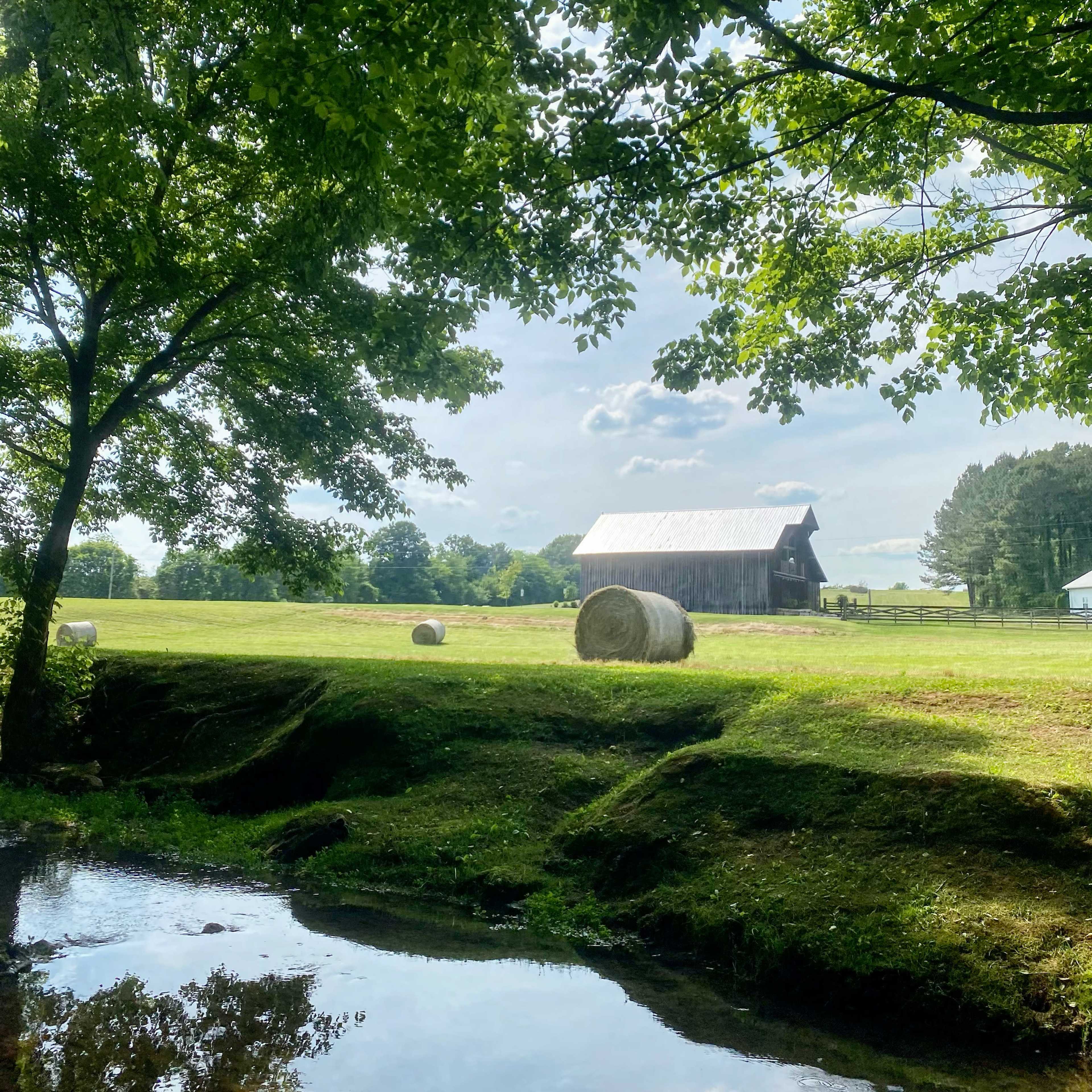 A barn stands in a lush green field beside a gently flowing stream, with hay bales scattered nearby.