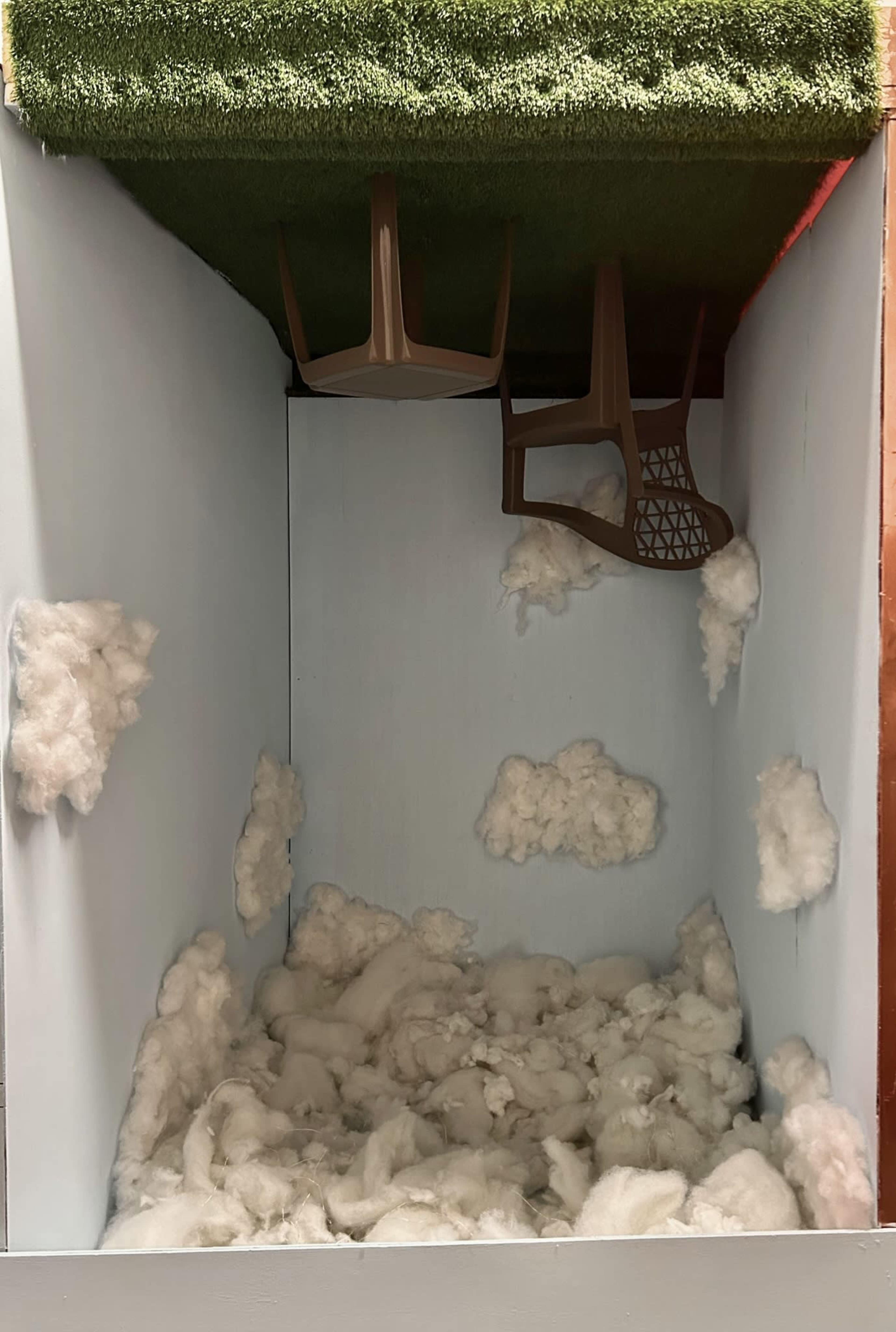 A diorama with a ceiling covered in green grass, a wooden chair inverted on the grass, and white cotton-like clouds scattered around the walls and floor.
