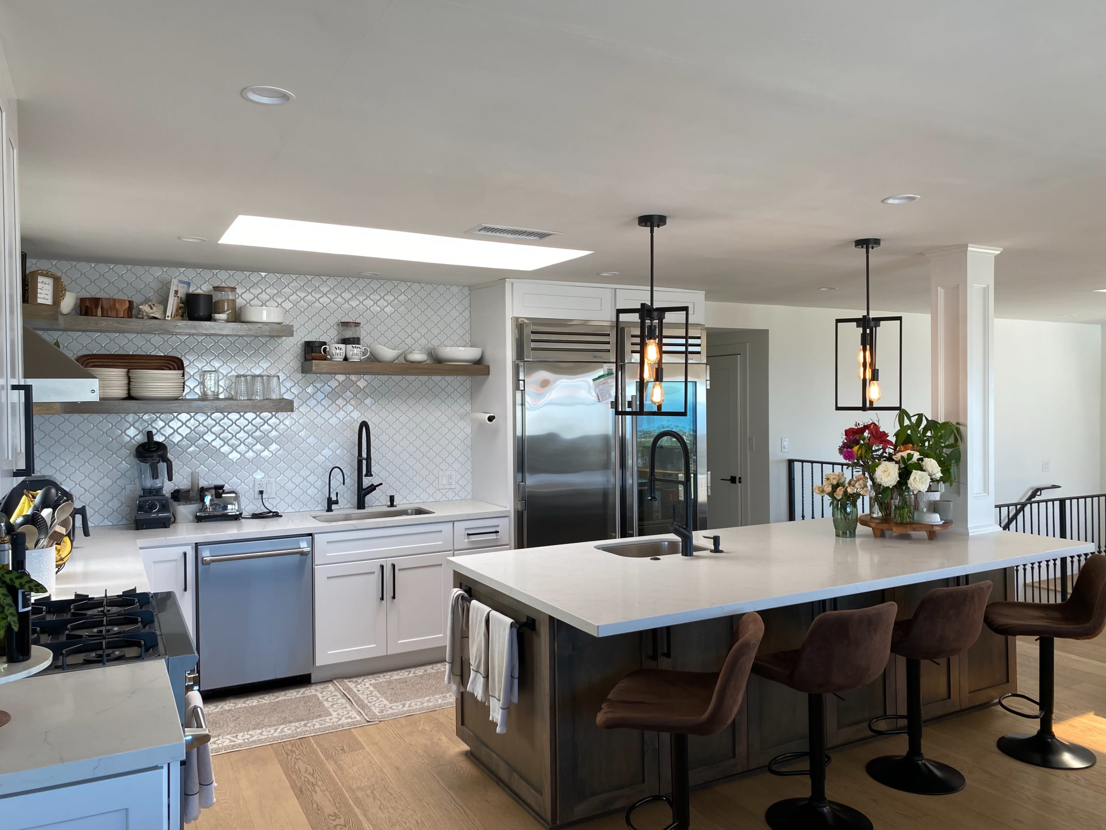 The kitchen features a large island with seating, modern appliances, white cabinetry, and decorative shelving with plants and dishware.
