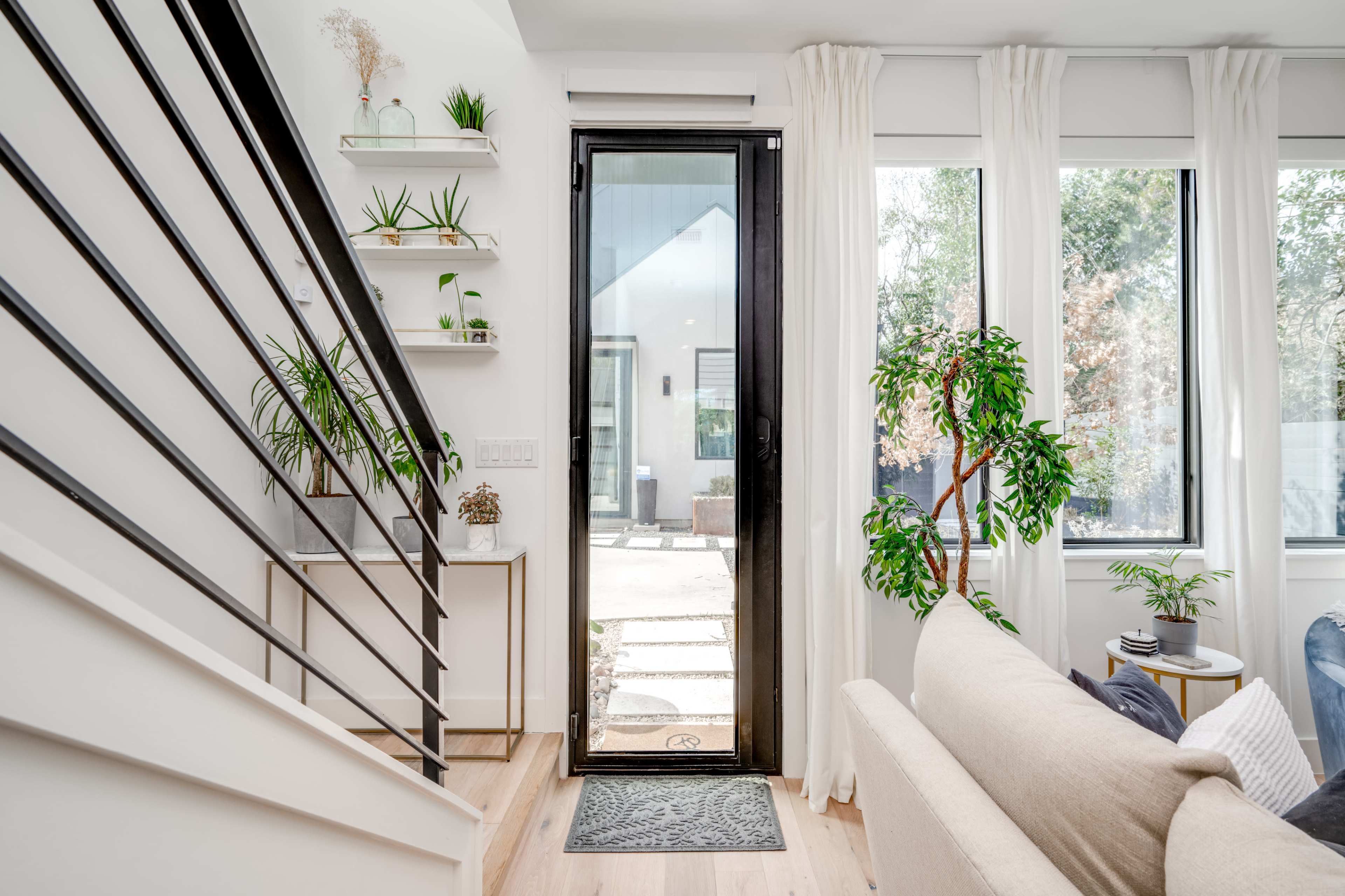 An entryway with a glass door, a staircase on the left, and greenery in planters near large windows.