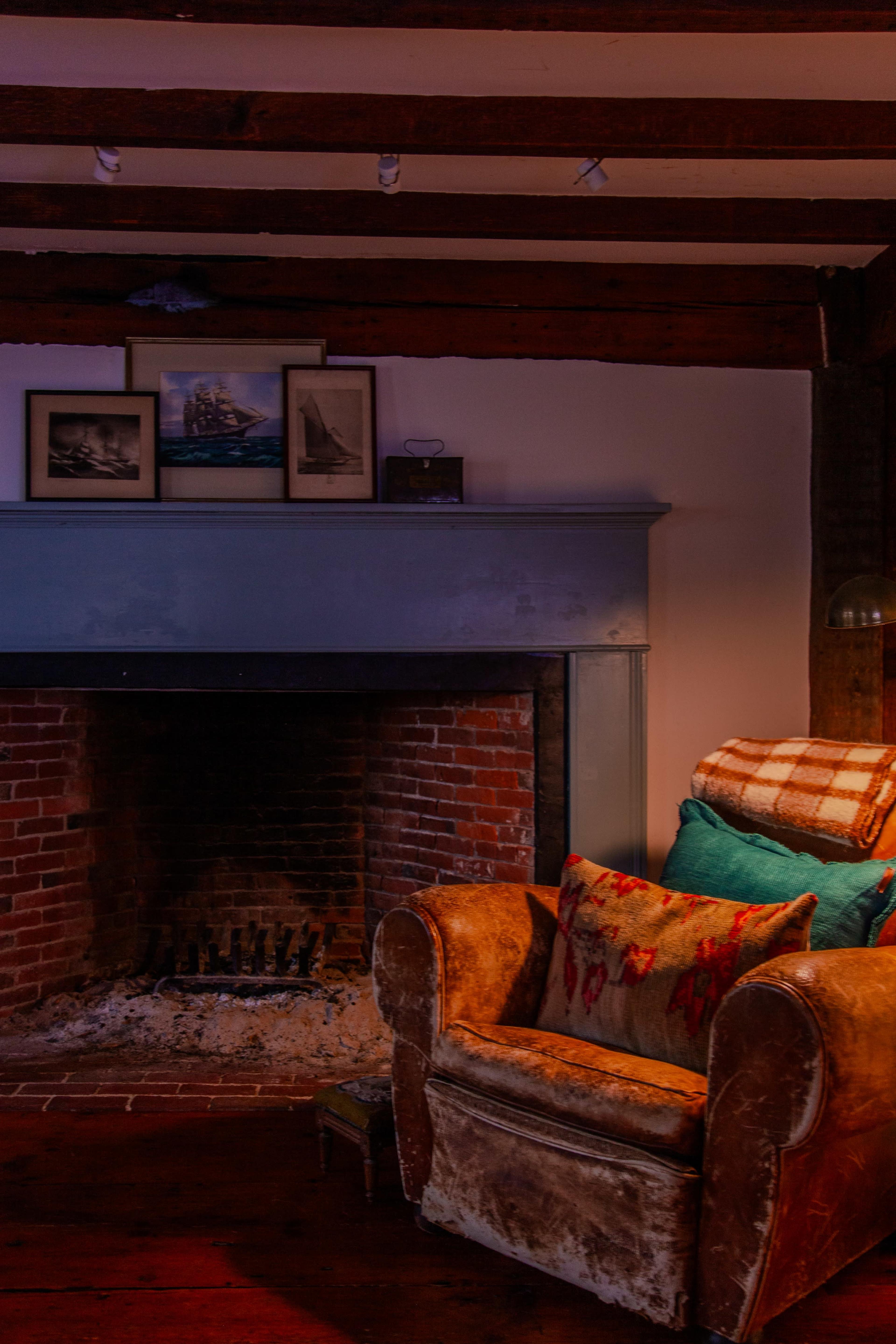 A worn armchair with decorative cushions sits beside a brick fireplace under wooden beams, featuring framed nautical images on the mantle.