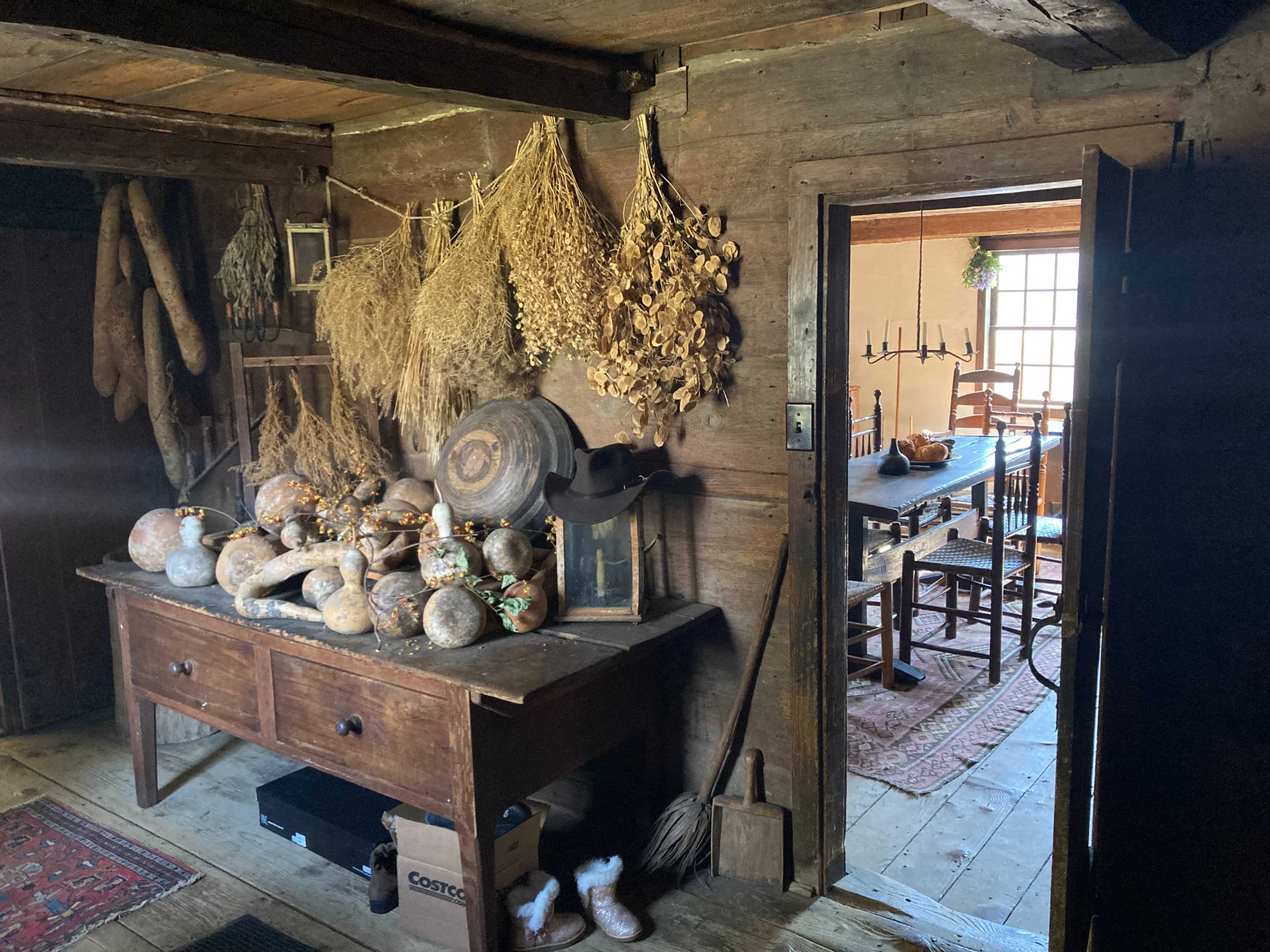 A rustic room with a wooden table covered in various gourds and surrounded by dried herbs, with a dining area visible in the background.