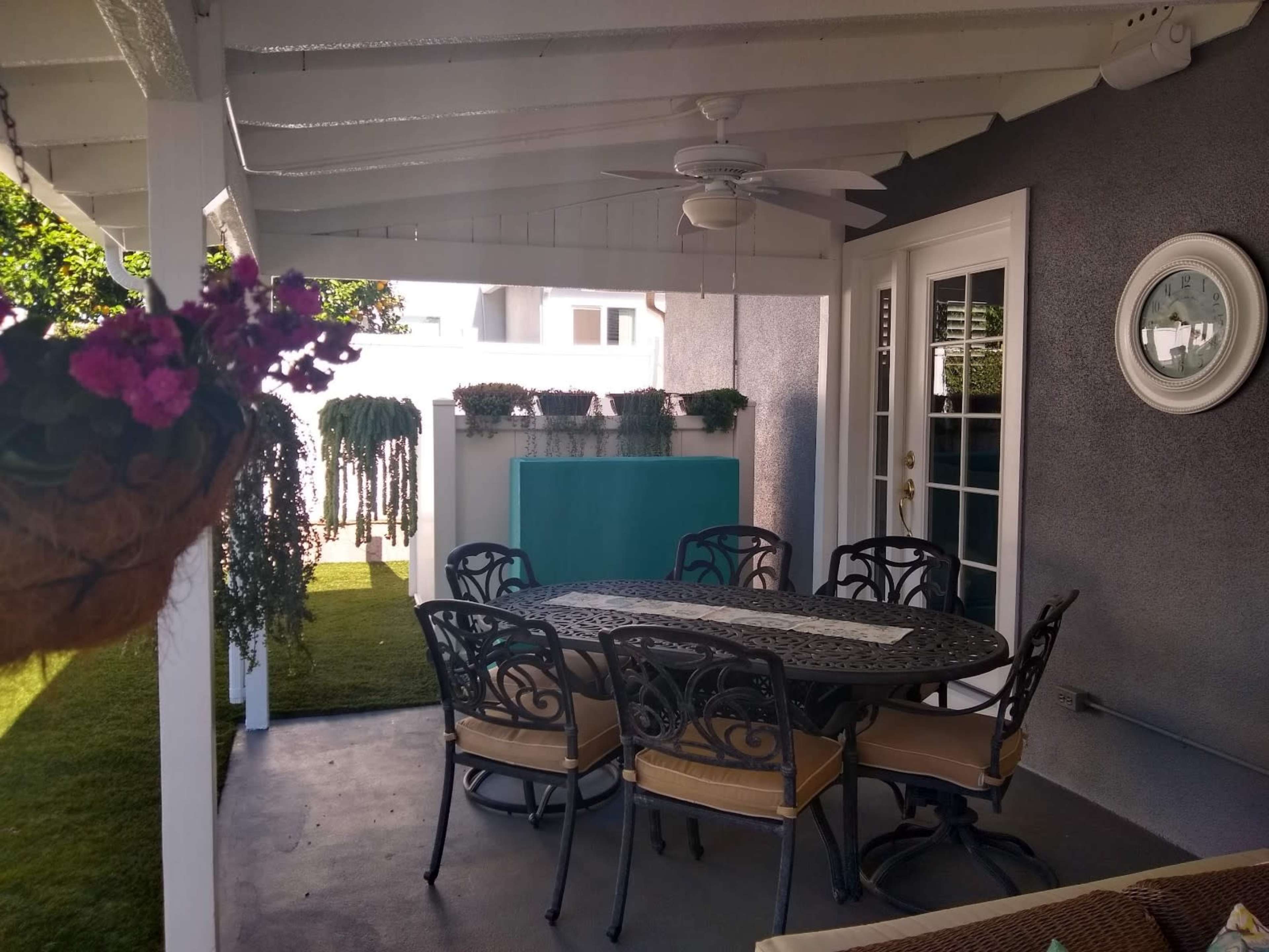 A covered patio area with a circular table and eight chairs, surrounded by potted plants and a wall-mounted clock.