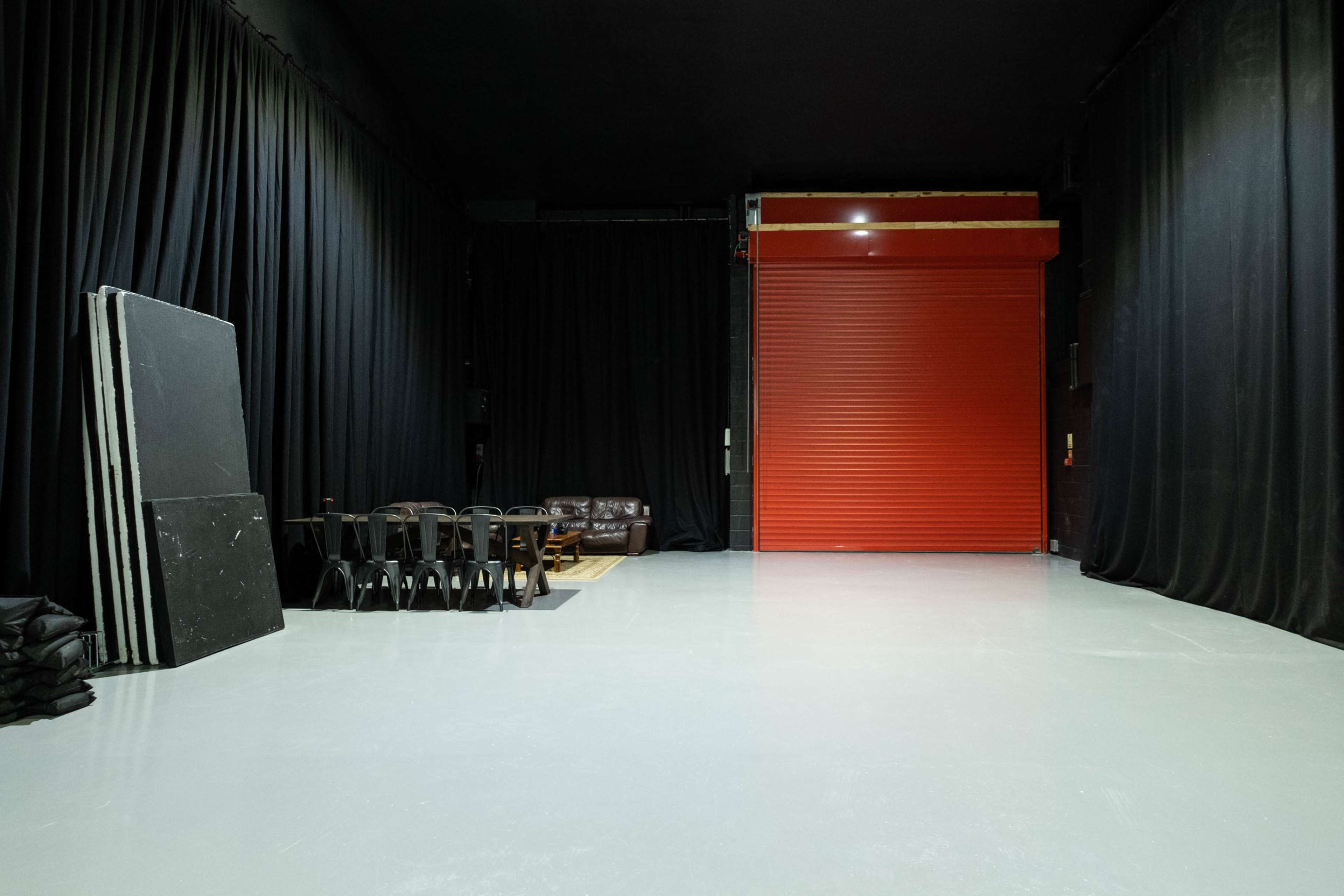 An empty studio space with black curtains, a red roll-up door, a brown couch, and a table with several chairs.