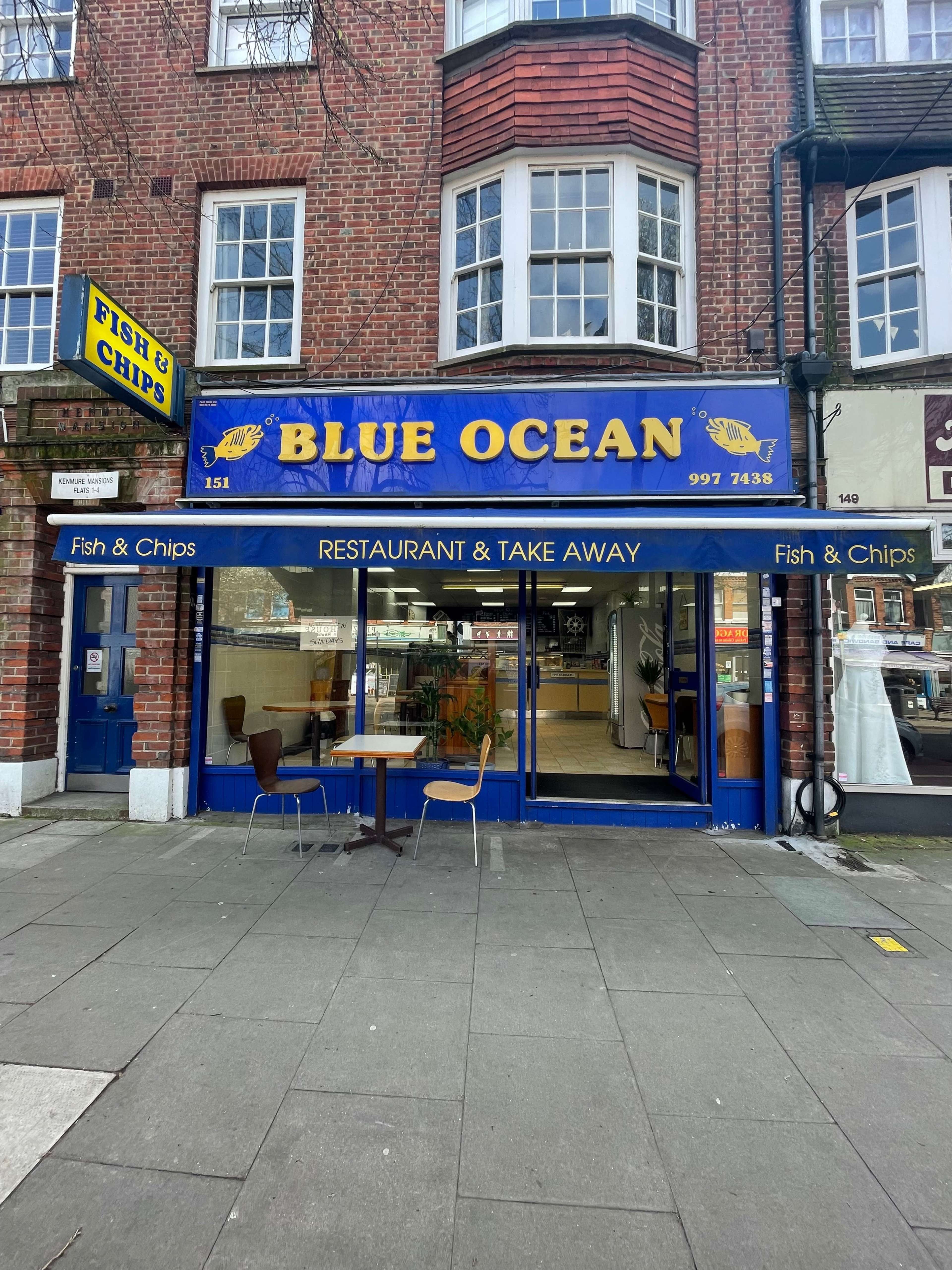 The exterior of a fish and chips restaurant called "Blue Ocean," featuring a blue sign and a large window with tables inside.