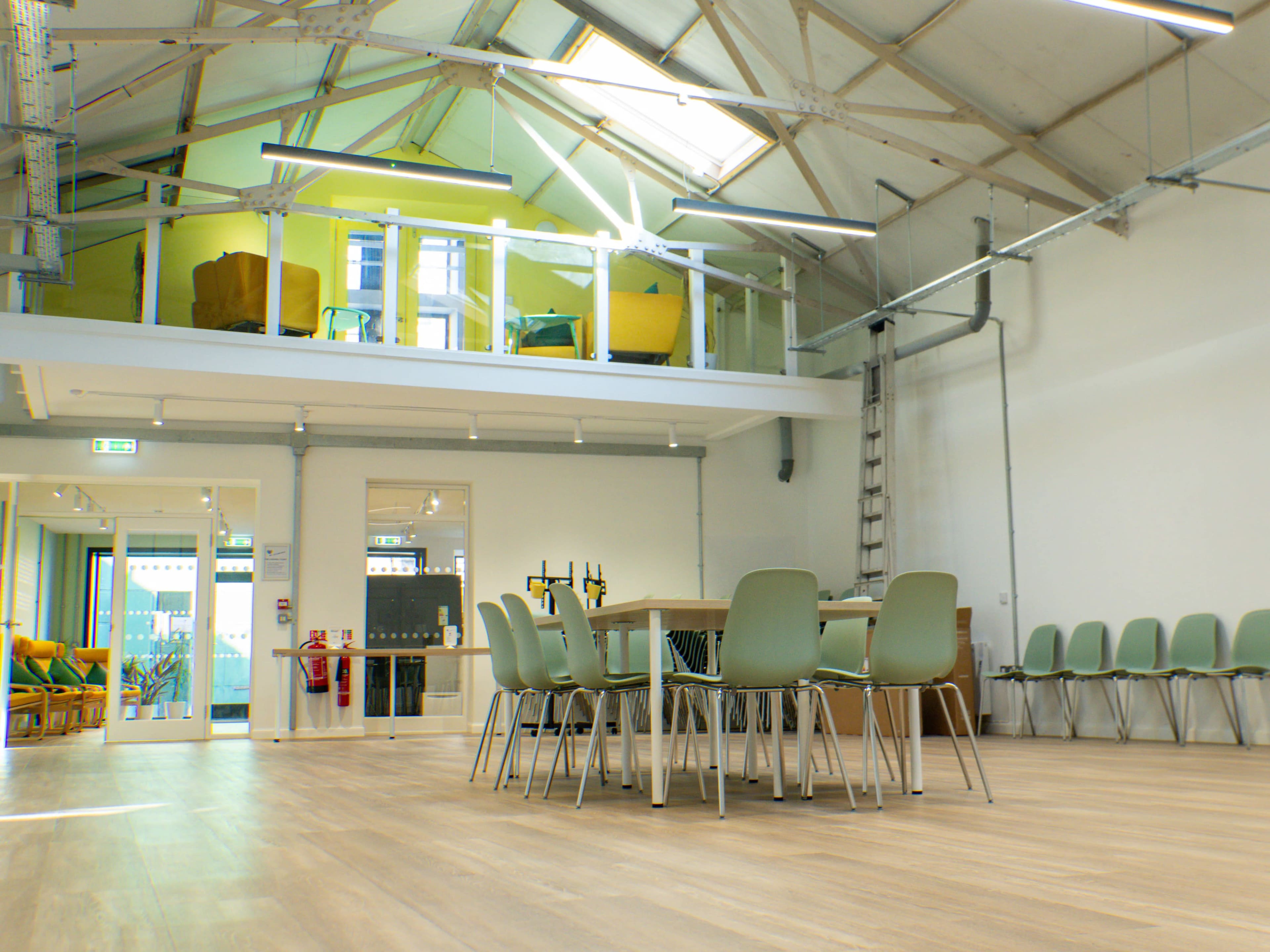 The image shows a spacious, modern interior of a building with light-colored wooden flooring, a central table surrounded by green chairs, and a lofted area with yellow seating.