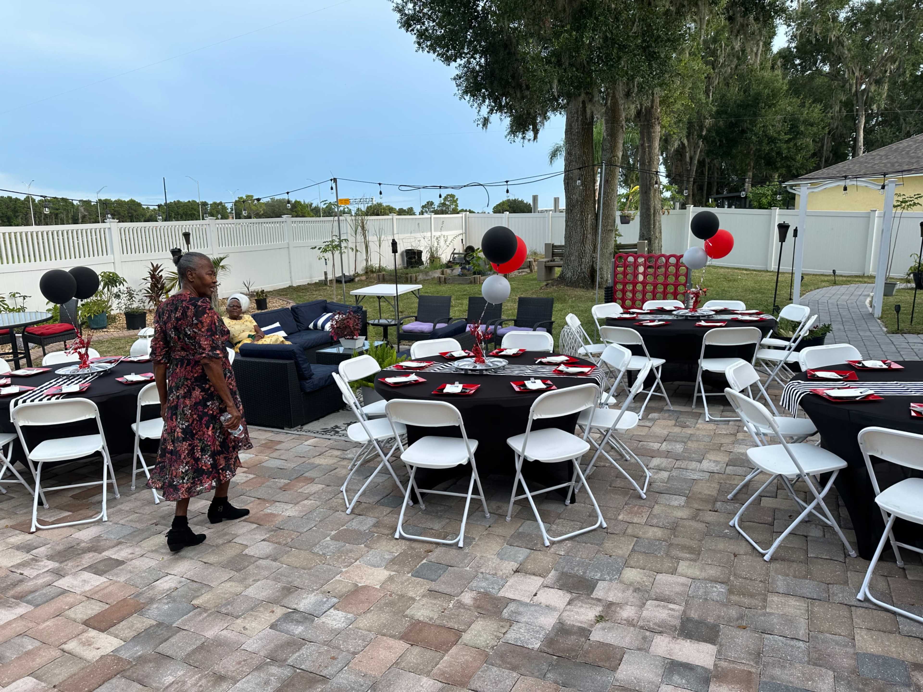 The outdoor space is arranged for a party with circular tables covered in black and red linens, balloons, and a seating area featuring a sofa.