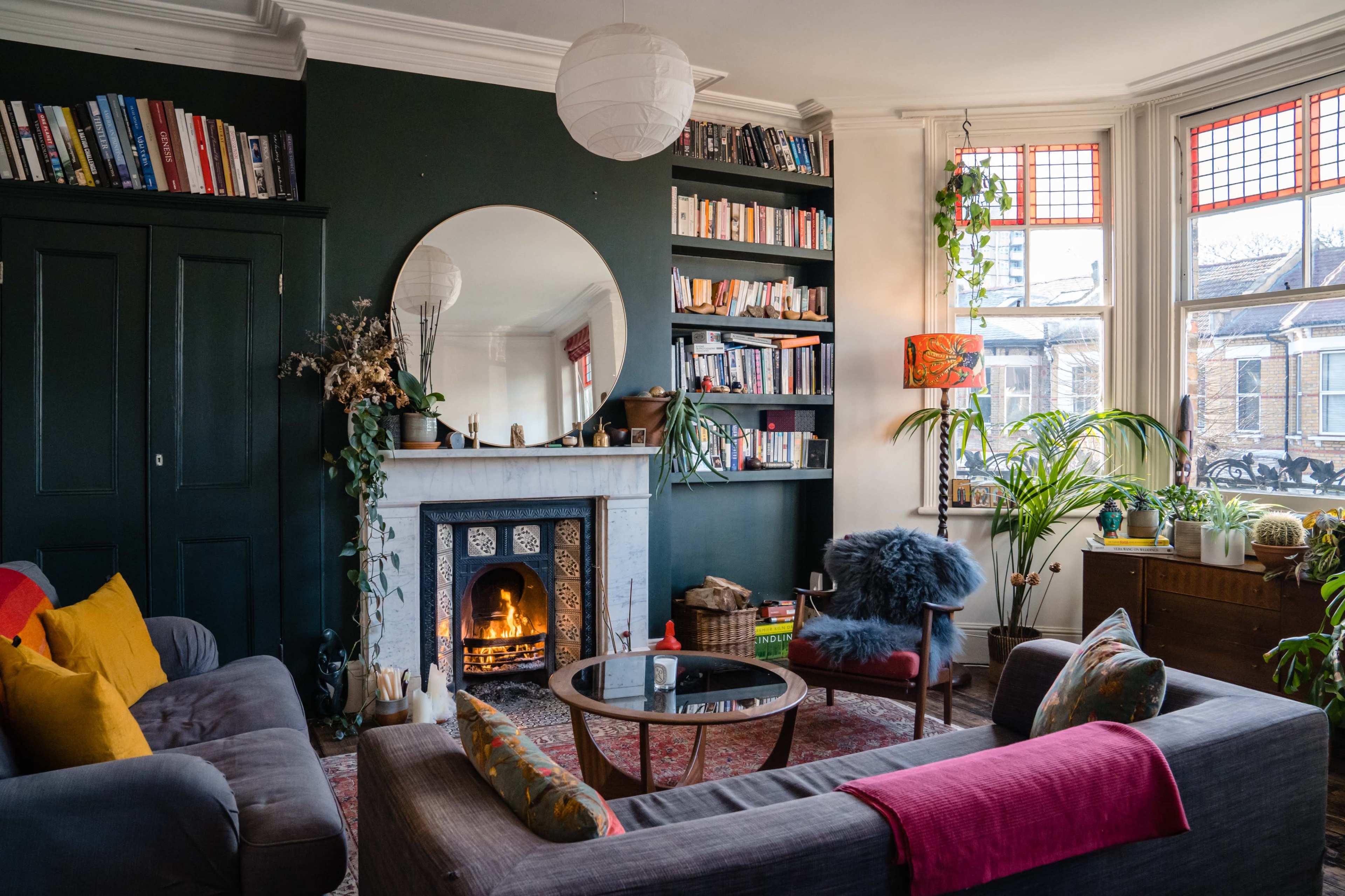 A cozy living room featuring a fireplace, bookshelves filled with books, and a large mirror reflecting a light fixture, with plants and decorative items present throughout the space.