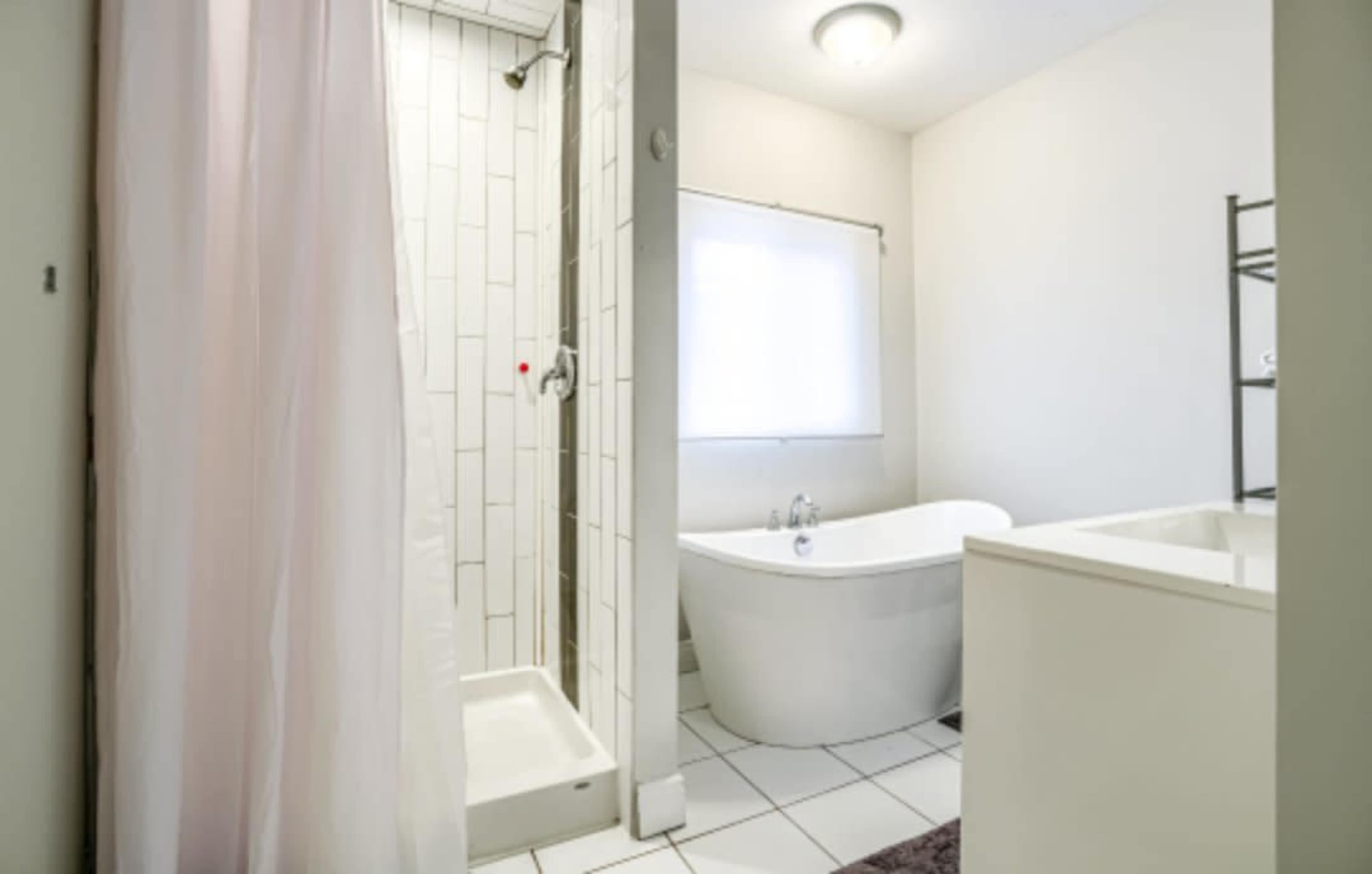 The image shows a bathroom featuring a shower with glass doors, a freestanding bathtub, and a window for natural light.
