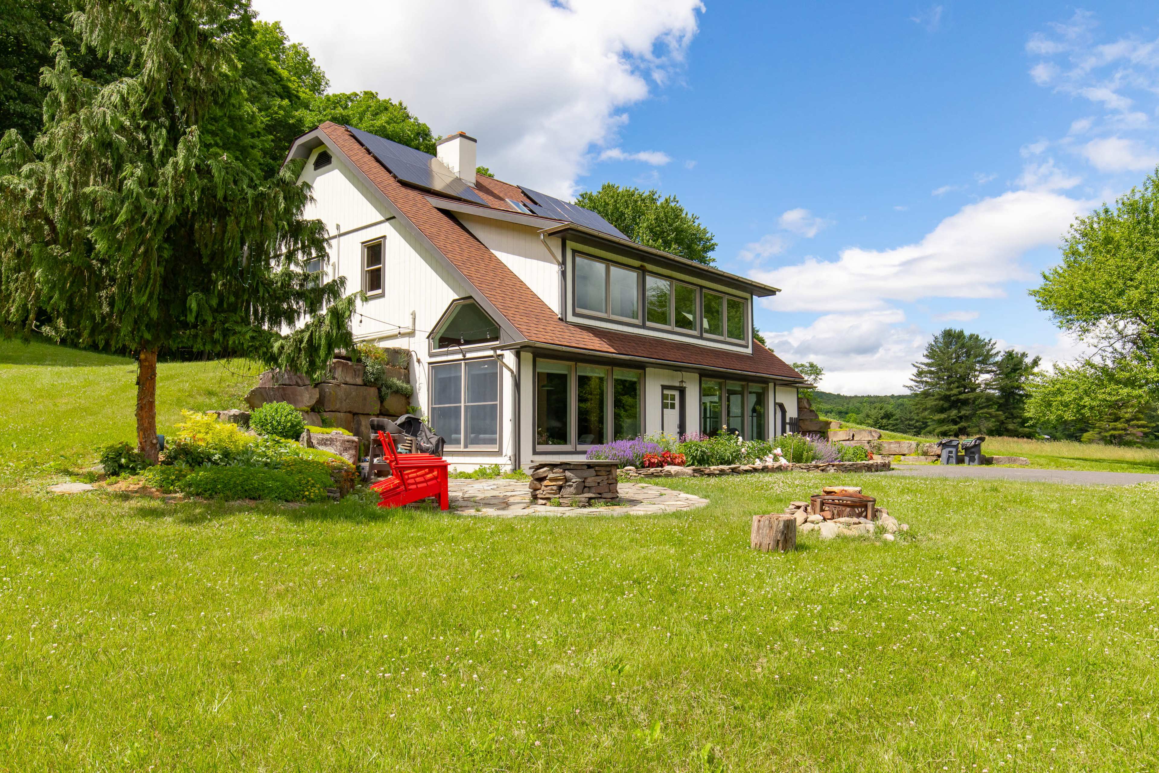 A modern, two-story house is situated on a grassy lawn with a stone patio and fire pit, surrounded by trees and landscaped gardens.