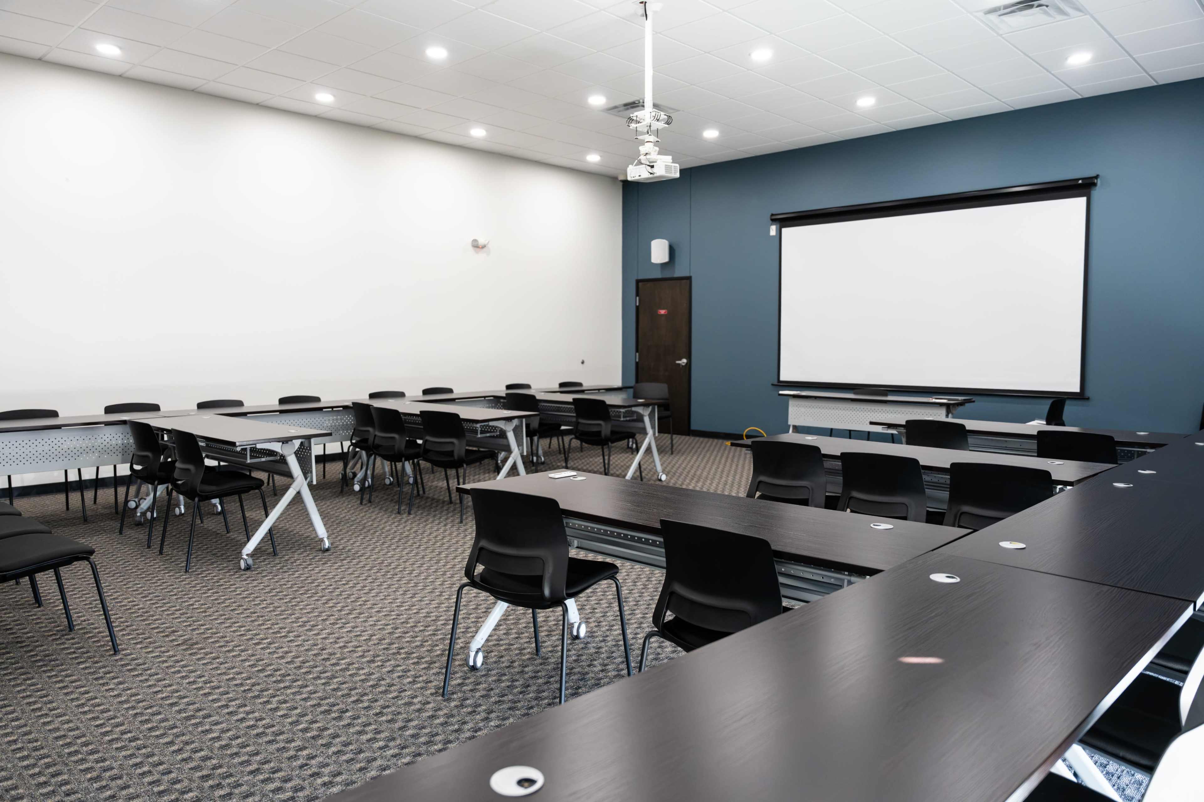 The image shows a modern classroom featuring rows of chairs and tables, with a projector screen and ceiling-mounted projector in a well-lit space.