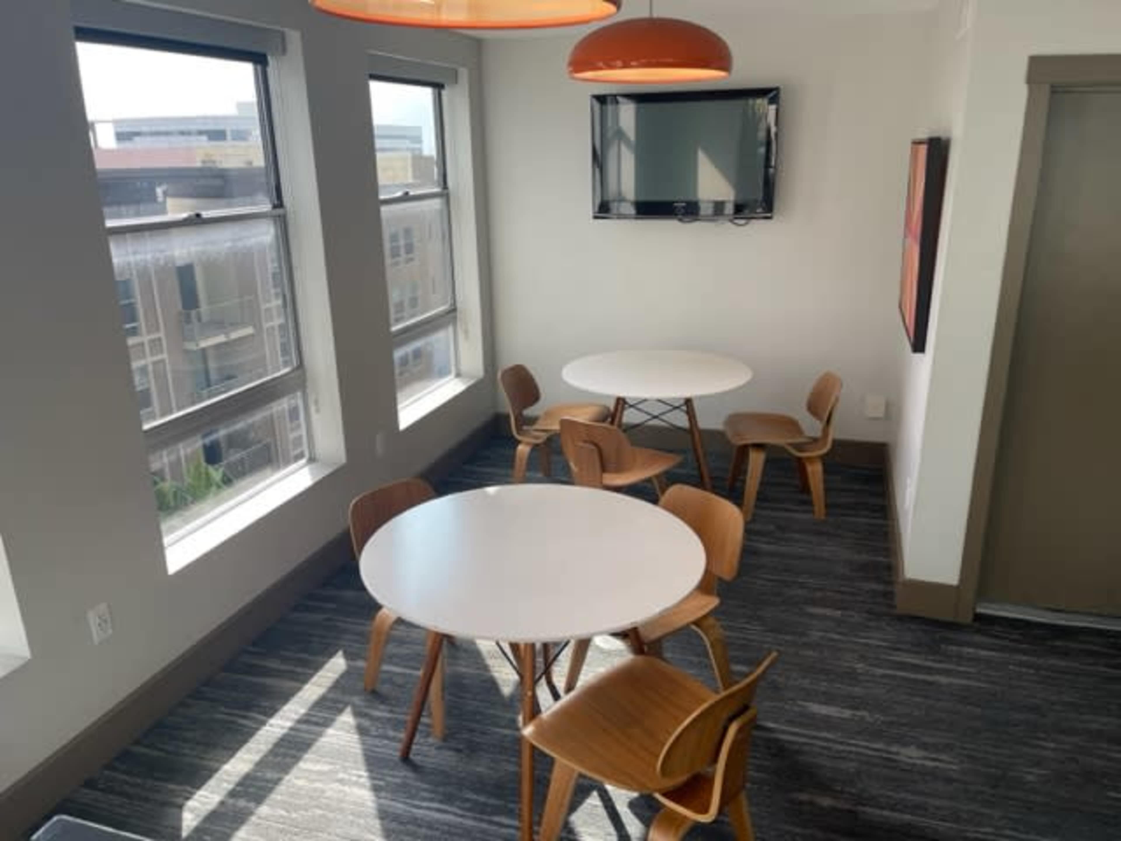 A well-lit room with two round white tables surrounded by wooden chairs and a wall-mounted TV.
