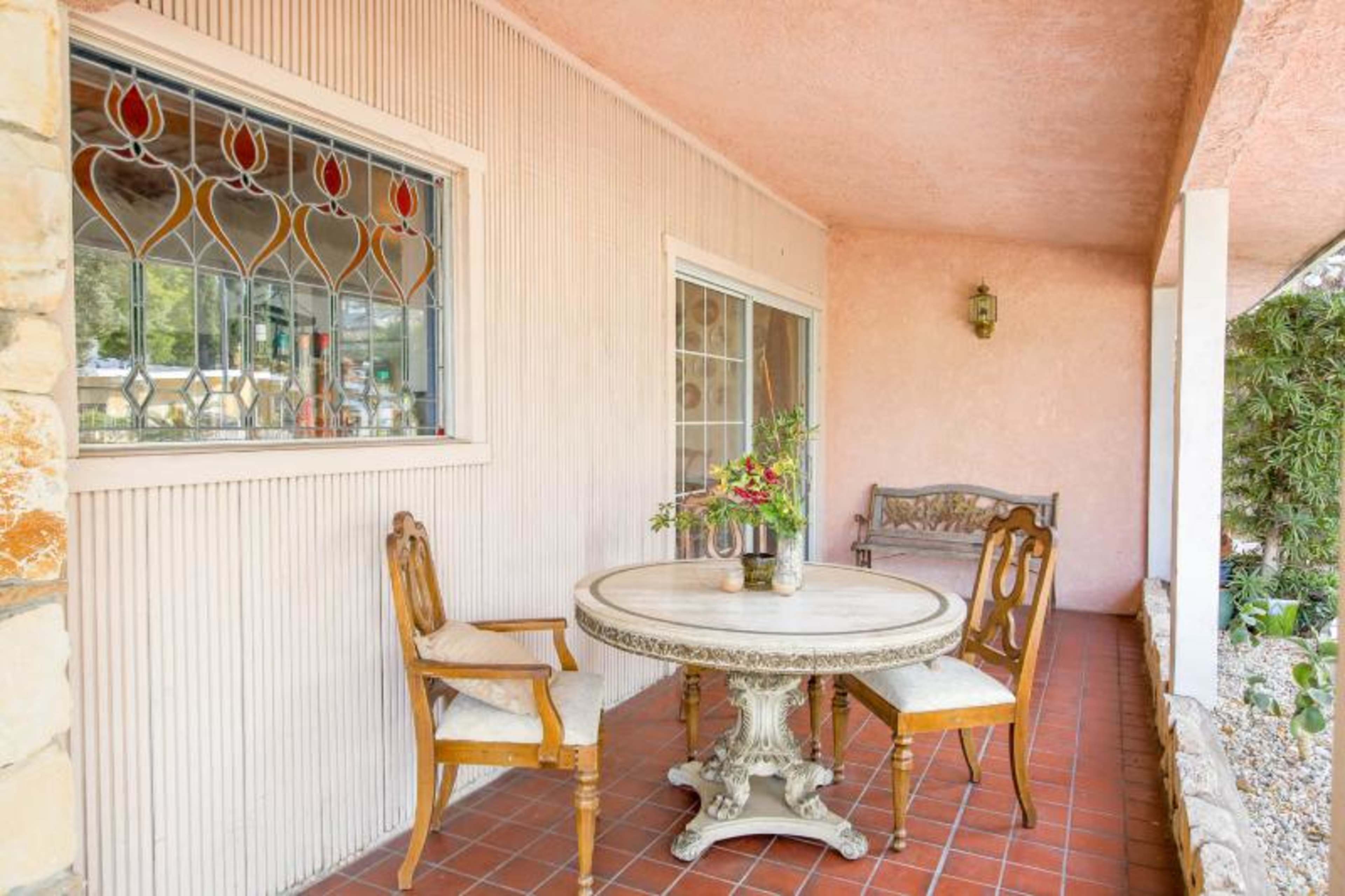A round table with floral decor and four chairs is set on a tiled patio adorned with a stone wall and a window decorated with stained glass.