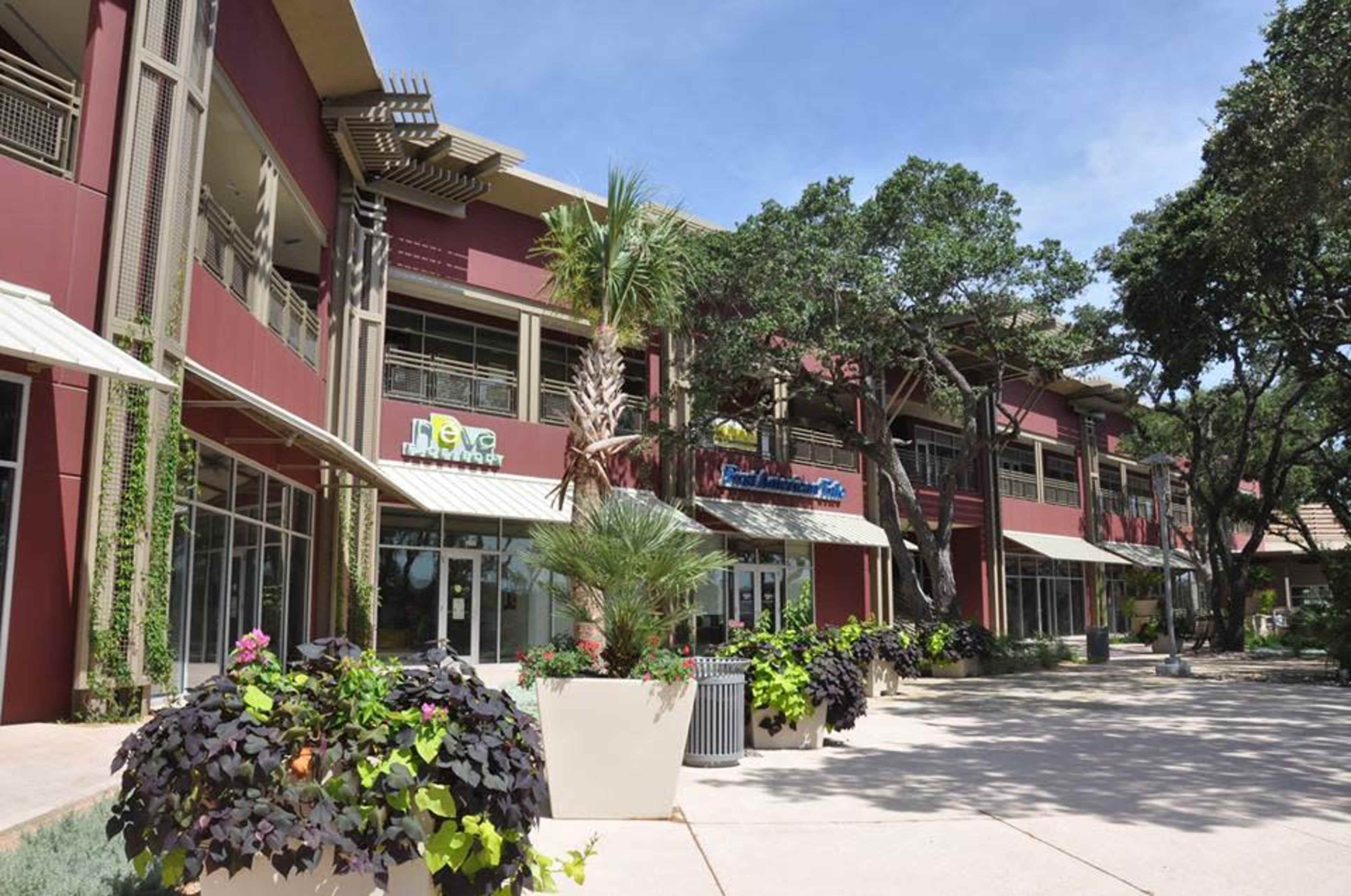 A commercial plaza with a row of businesses, surrounded by greenery and large planters.