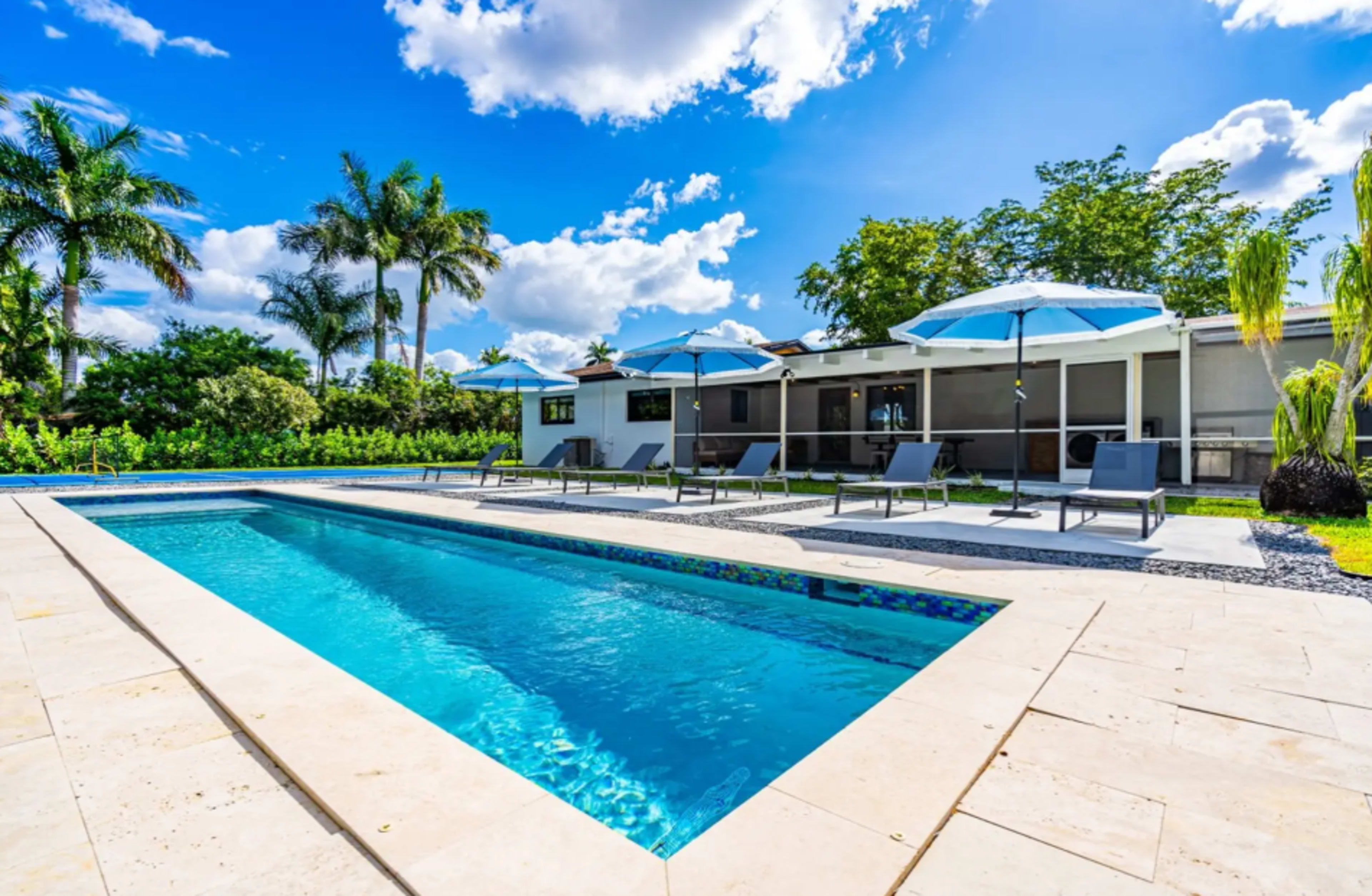The image shows a modern outdoor swimming pool surrounded by lounge chairs and umbrellas, with a house in the background and palm trees under a blue sky.