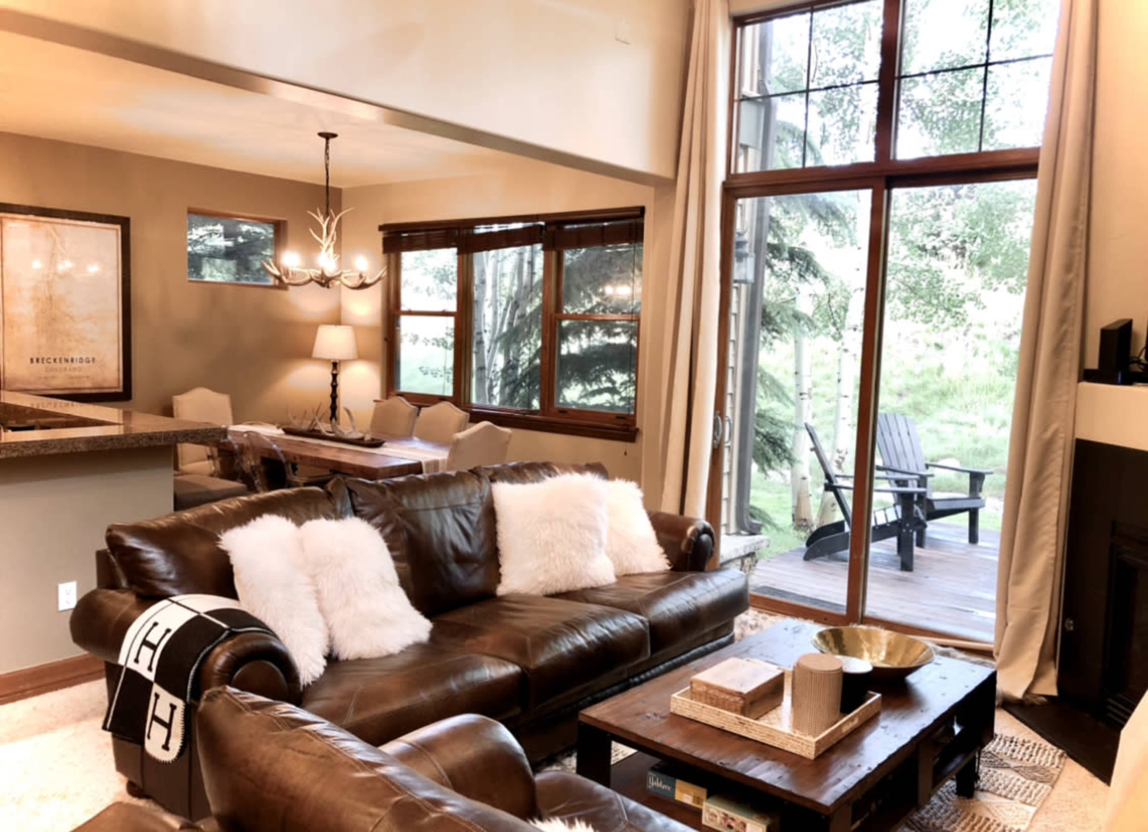 A cozy living room features a brown leather sofa with white throw pillows, a wooden coffee table, and large windows that open to a deck overlooking greenery.