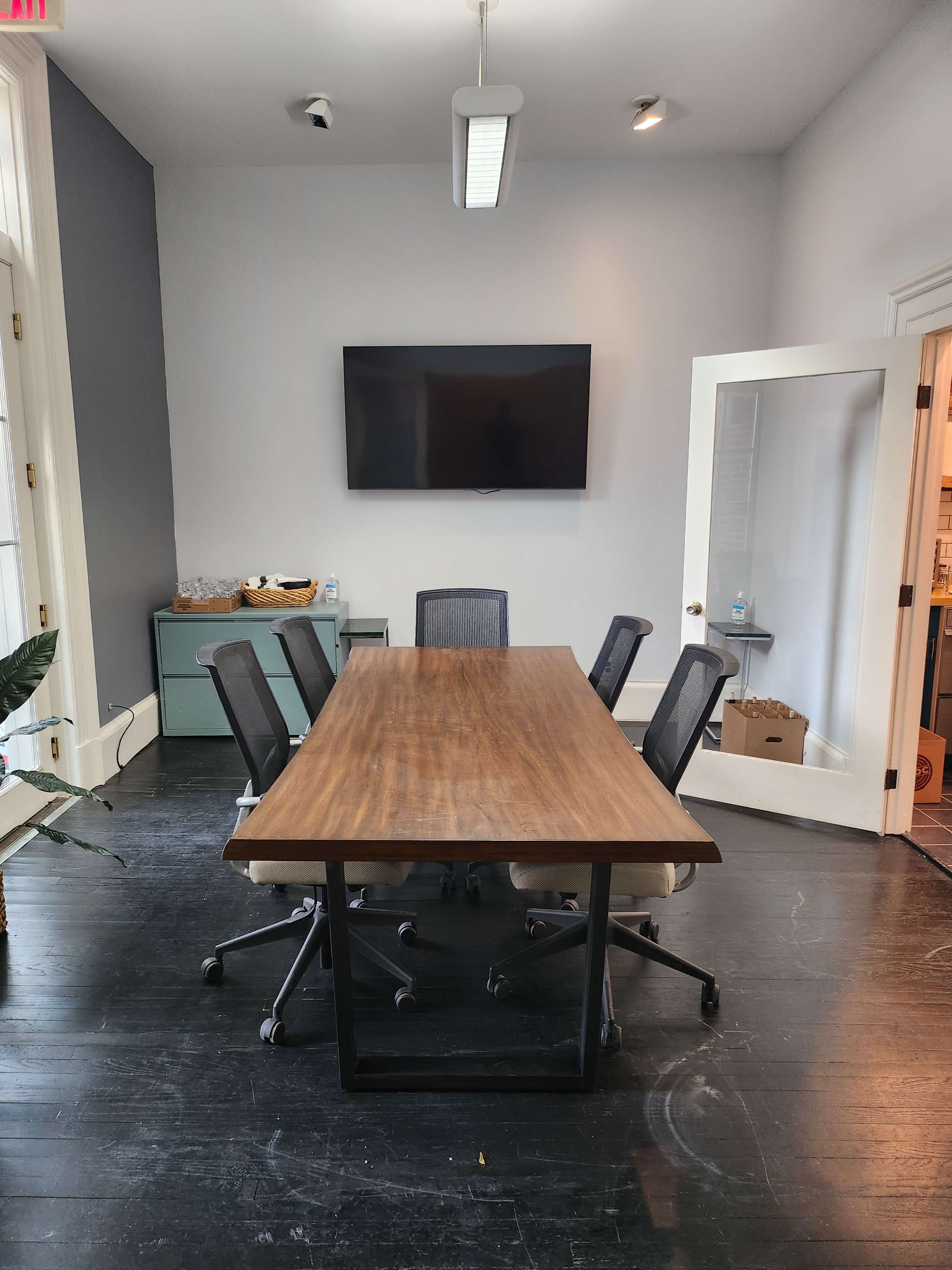 A modern conference room featuring a long wooden table, six black office chairs, a large television mounted on the wall, and a glass door leading to another room.