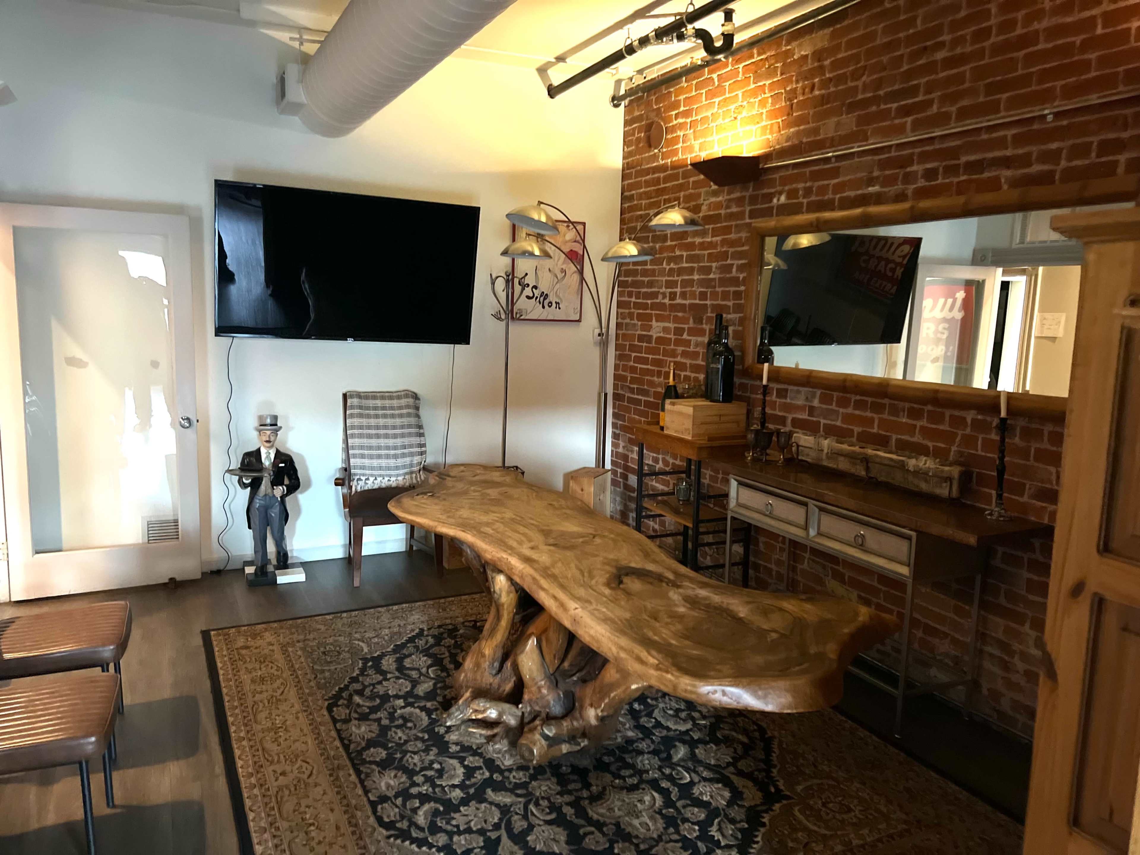 A room with a large wooden table, a vintage-style chair, a flat-screen TV mounted on a brick wall, and a mirror above a console table.