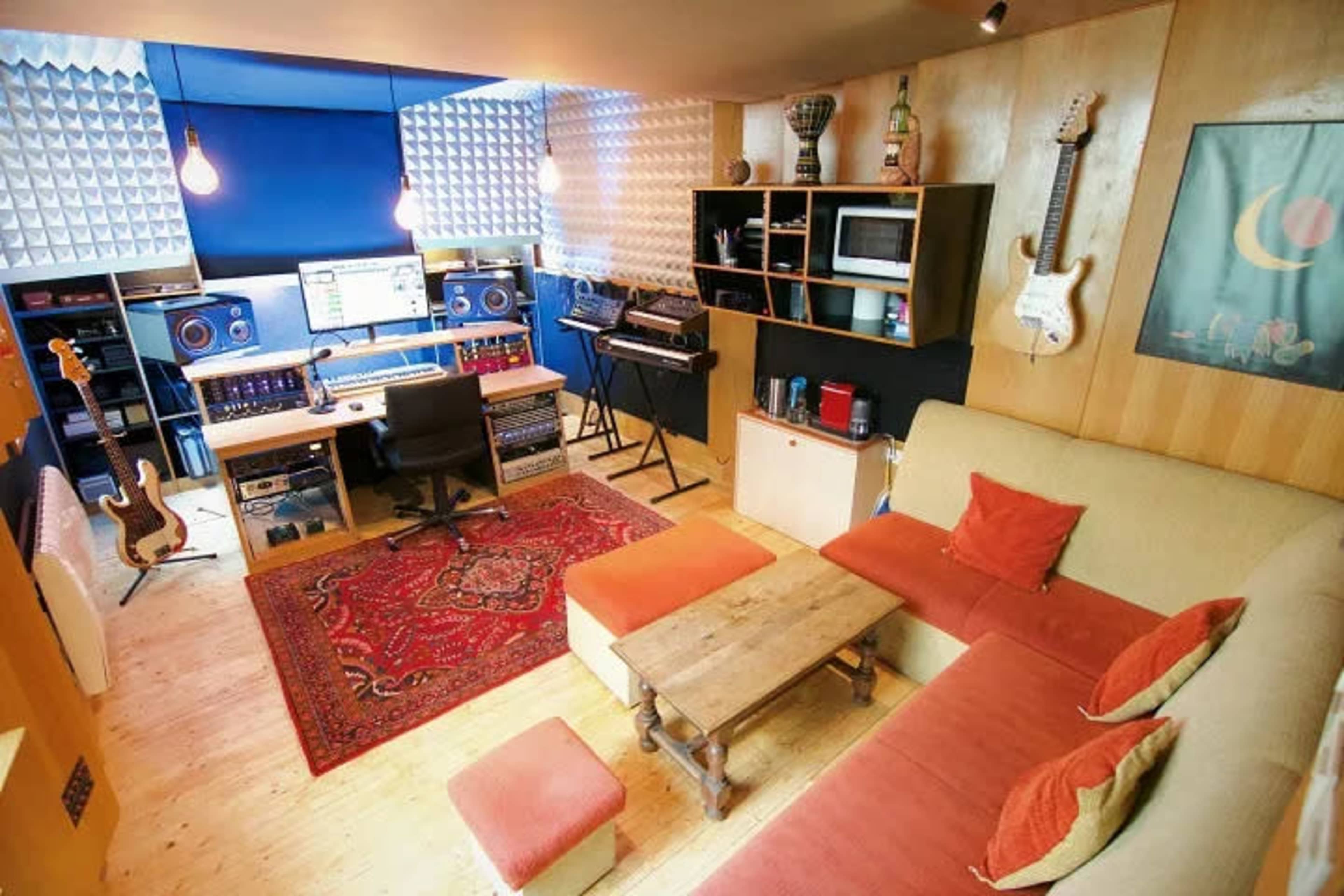 The image shows a small music studio with soundproofing panels, a computer setup with audio equipment, a couch with orange pillows, and various musical instruments displayed on the walls.